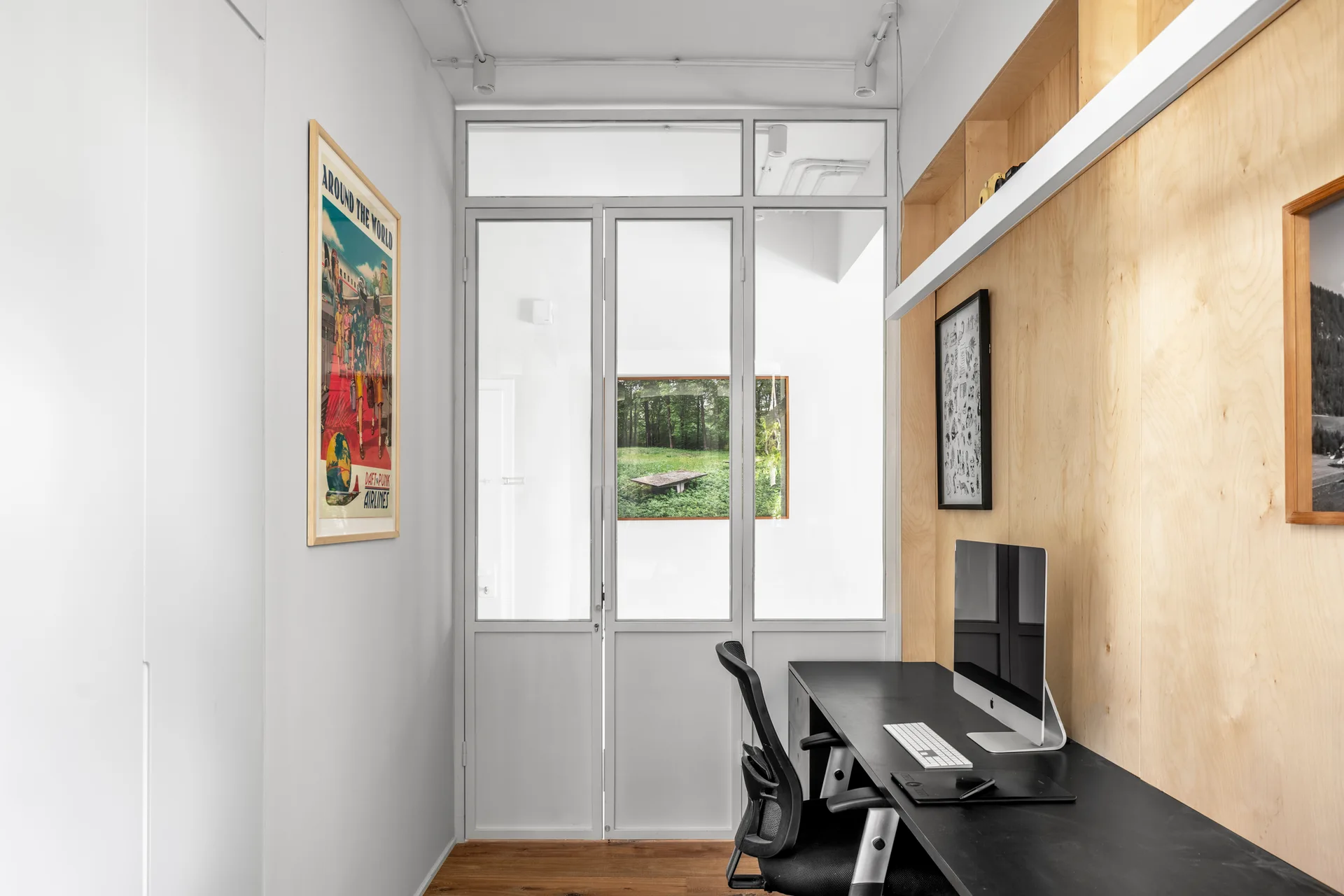 A narrow home office workspace featuring a black desk against a light wood paneled wall, separated by a grey framed glass partition door.