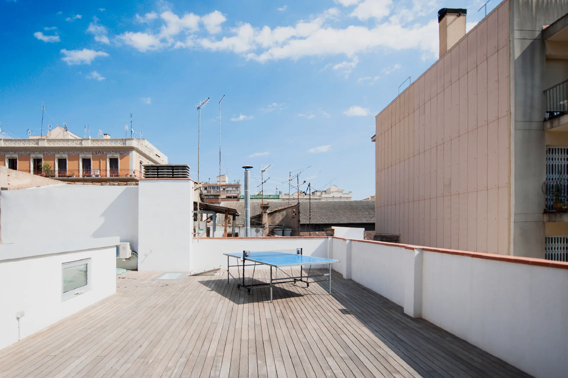 A spacious rooftop terrace features natural wood plank decking, tall white parapet walls, and a blue ping pong table in the center.