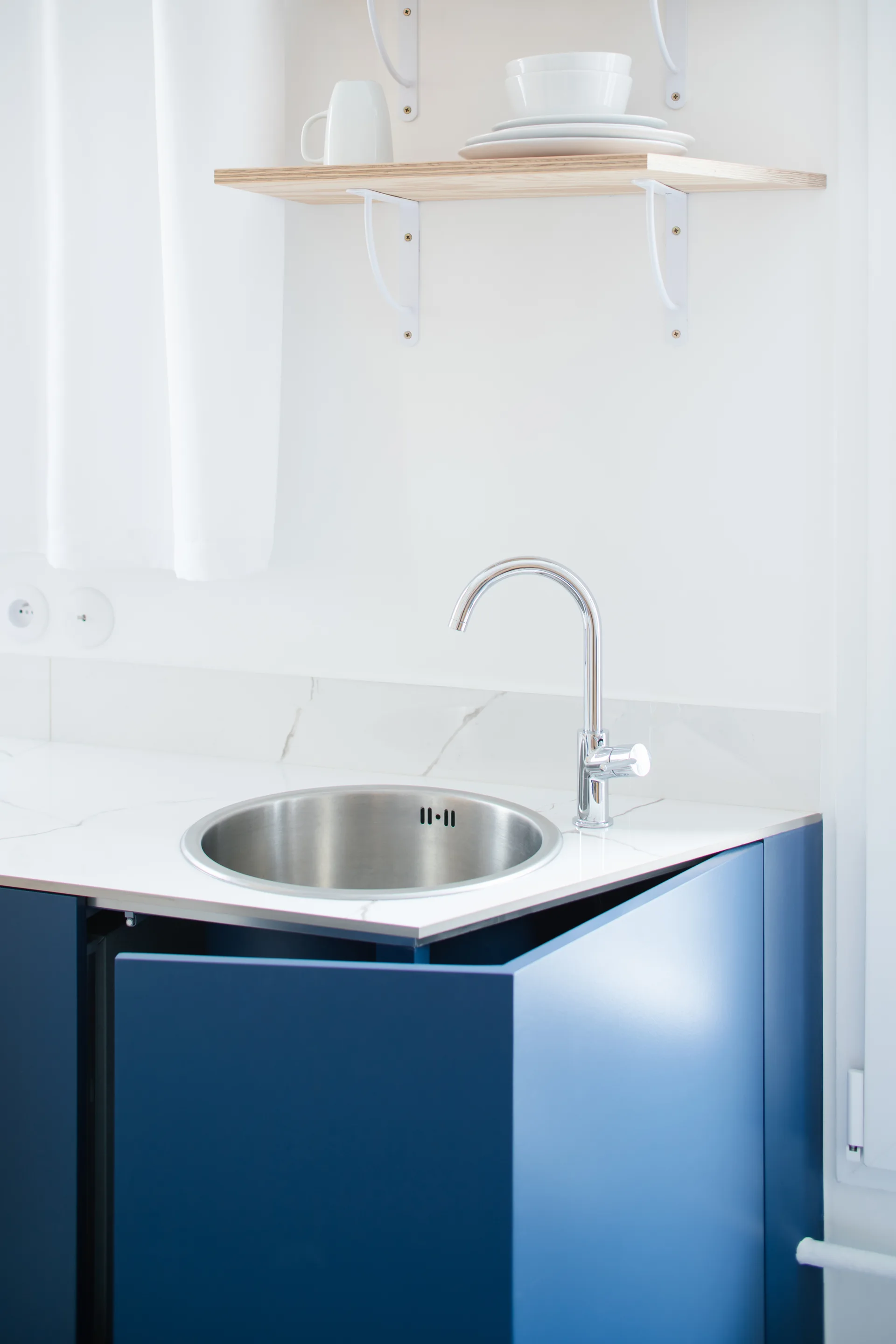 A close-up of a minimalist kitchen counter with blue matte cabinets, a round stainless steel sink, a chrome faucet, and a light wood wall shelf.