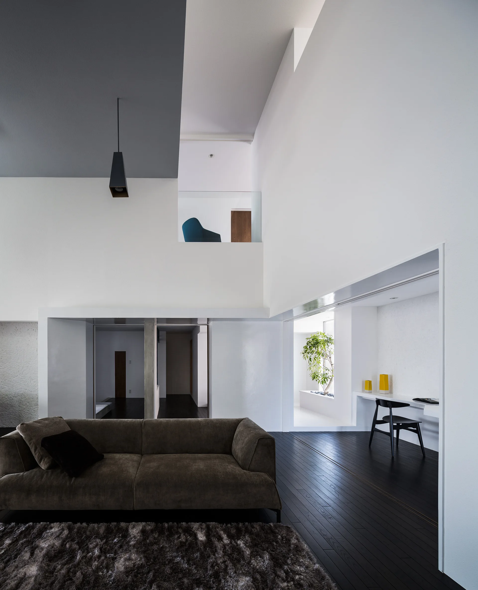 A minimalist double-height living room featuring a brown sofa, dark wood floors, a mezzanine level, and a built-in white desk in an alcove.