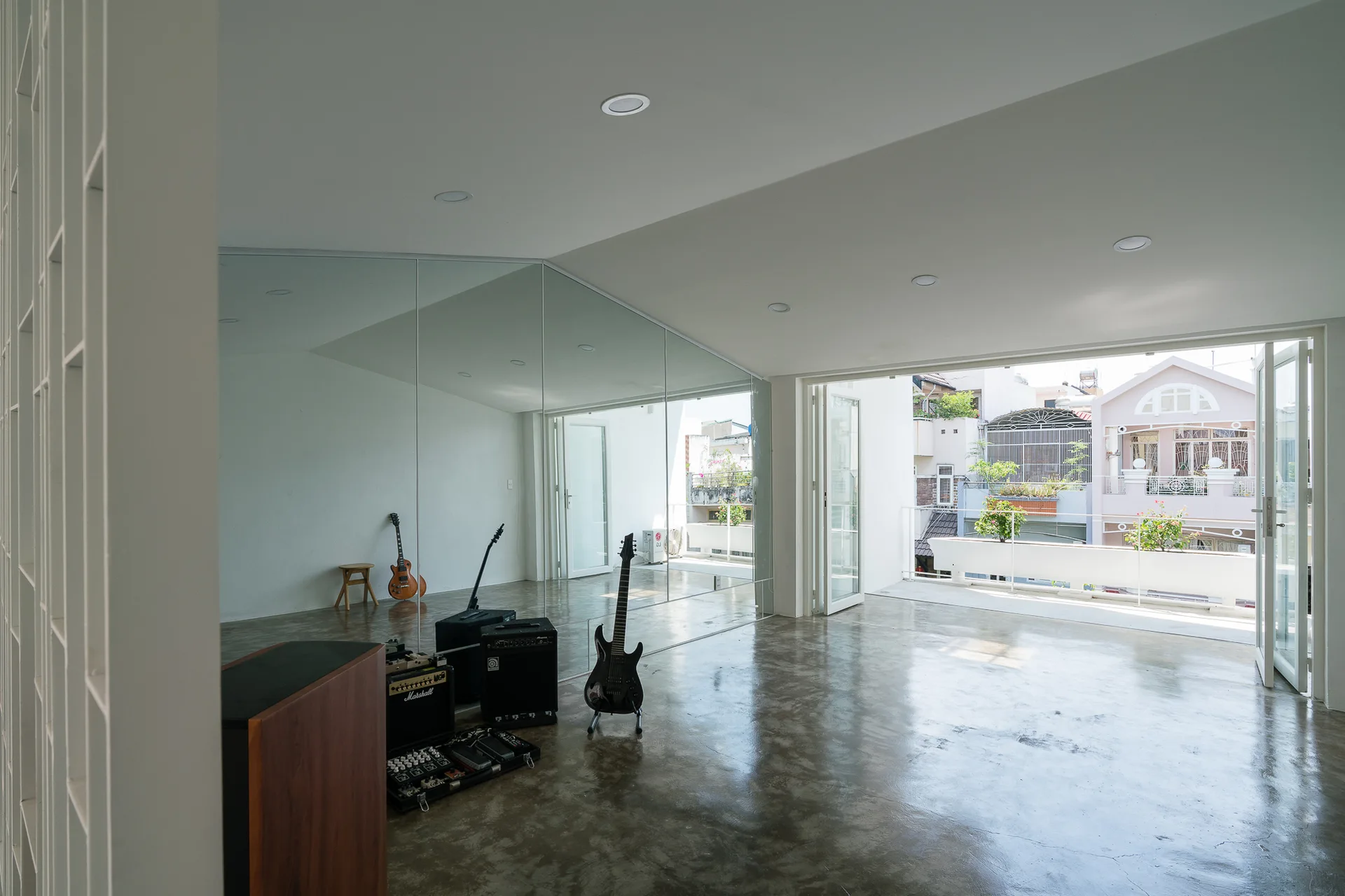 A minimalist home music studio features polished concrete flooring, guitars with amplifiers, a large mirrored wall, and bi-fold doors opening to a balcony.