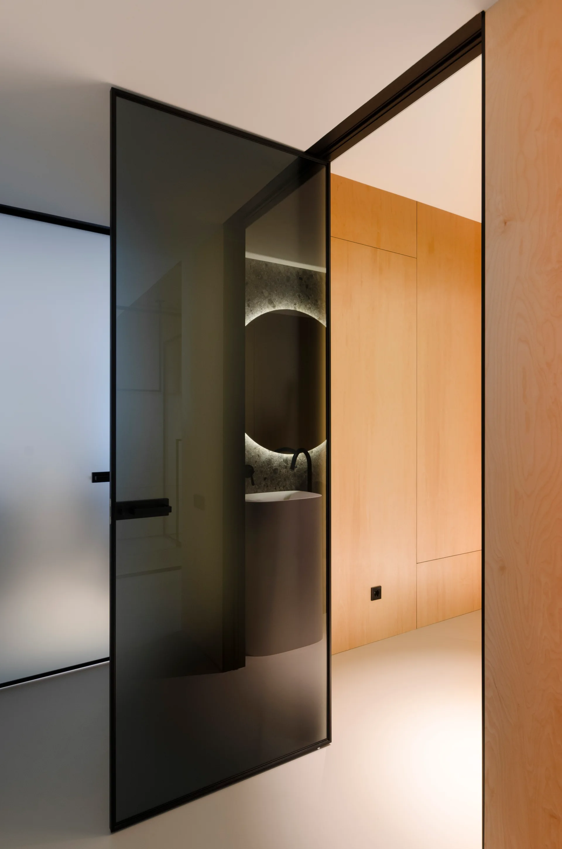 A modern bathroom with a cylindrical pedestal sink and a round backlit mirror is seen through an open smoked glass pivot door set in light wood paneling.