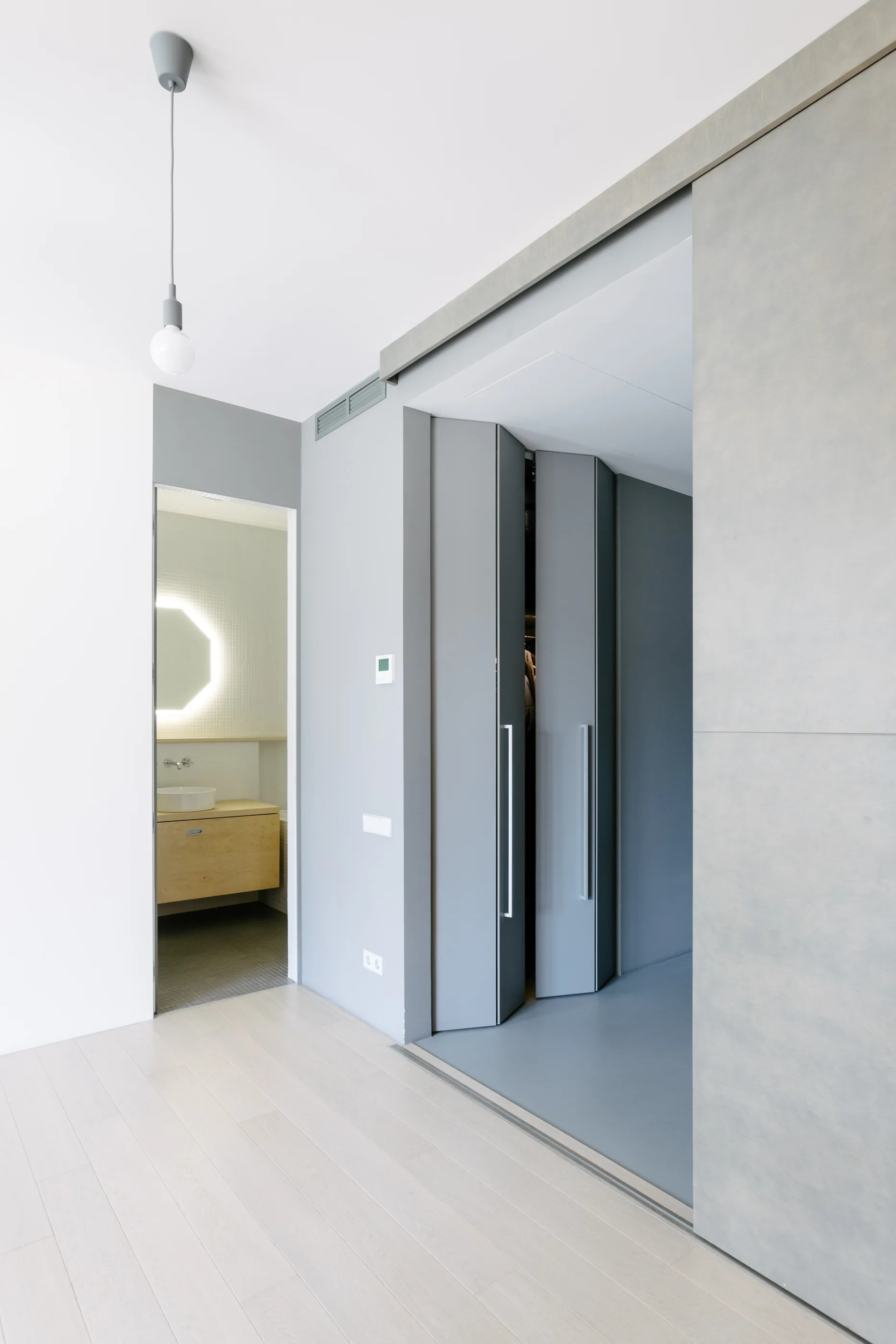 A grey hallway features bi-fold closet doors, light wood flooring, and a view into a bathroom with a backlit mirror and floating vanity.