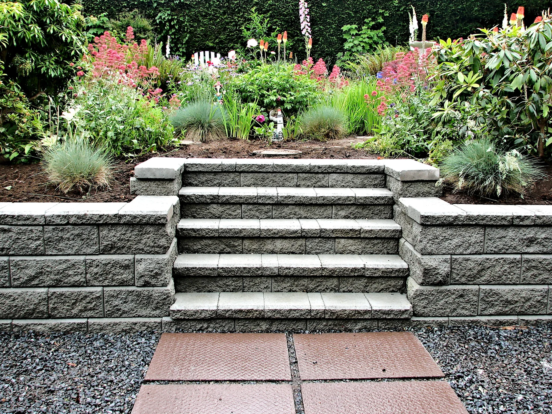 A textured grey stone retaining wall with central steps leads from a gravel patio up to a lush garden bed filled with green plants and pink flowers.