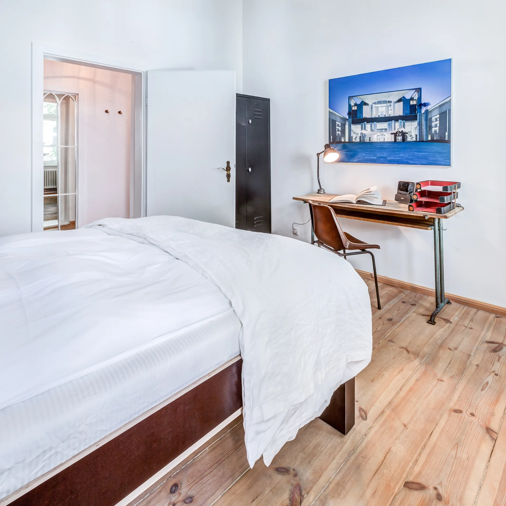 A bright bedroom displays a white bed, an industrial wooden desk with a leather chair, and a tall metal locker on wide wood floorboards.