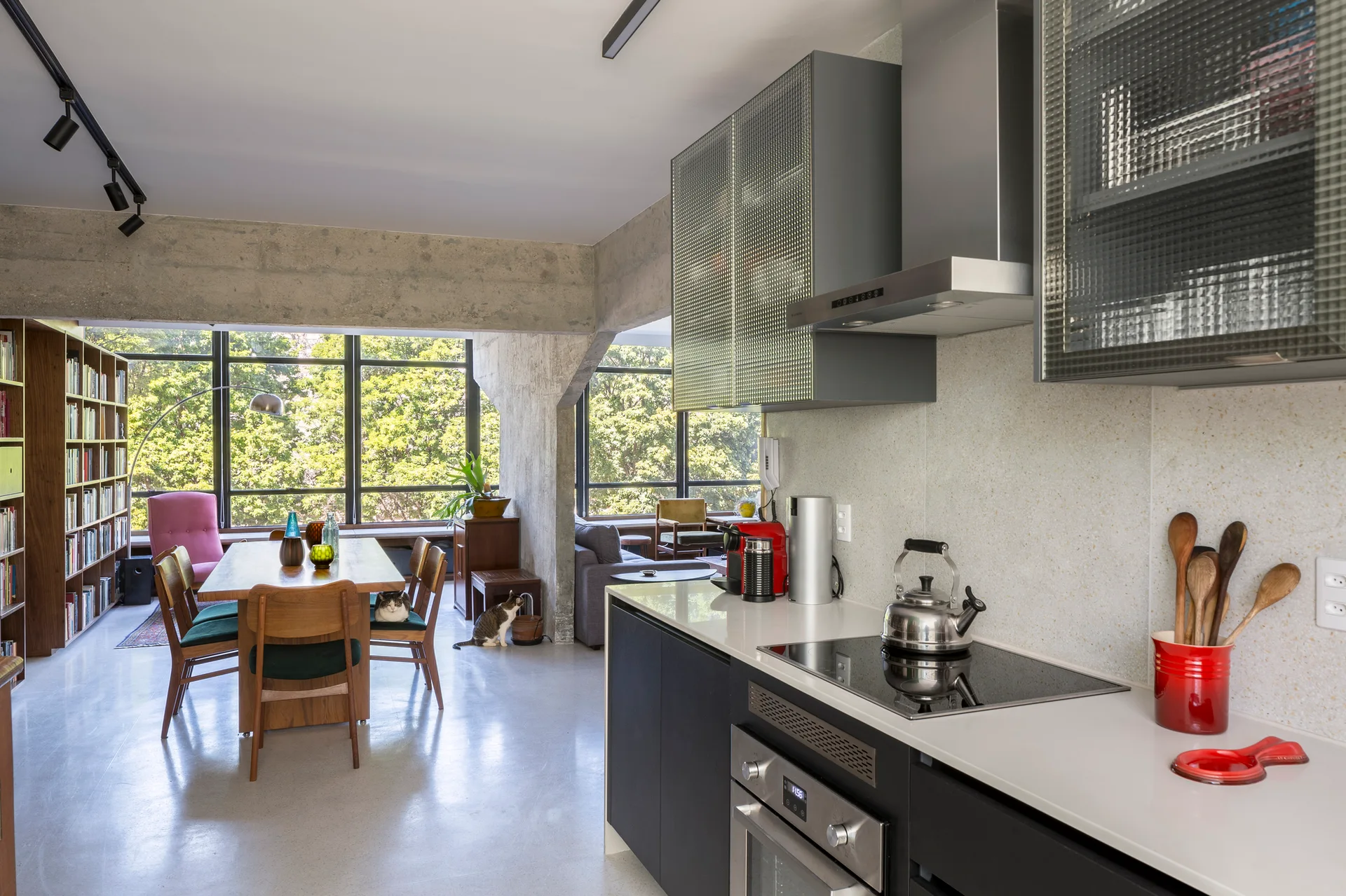An open-concept kitchen and dining area featuring dark cabinetry, textured glass upper cabinets, an exposed concrete support column, and a wooden dining set.