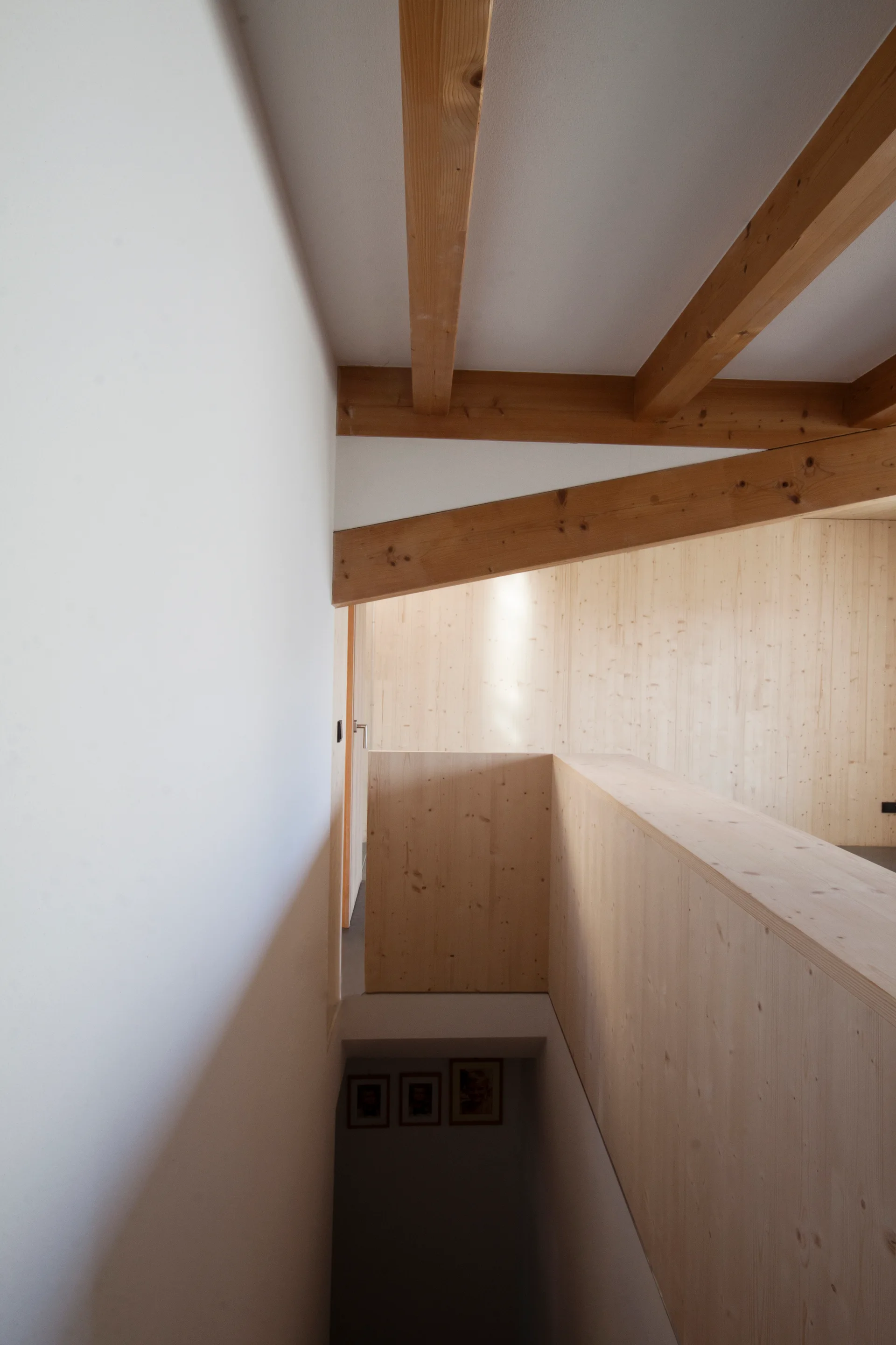 A modern stairwell with light wood paneled walls, a solid wood balustrade, and exposed timber ceiling beams next to a white wall.