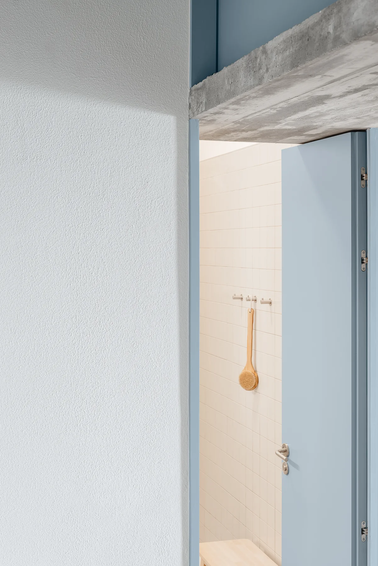 An open dusty blue door reveals a minimalist bathroom with off-white tiled walls, metal wall hooks, and an exposed raw concrete beam overhead.