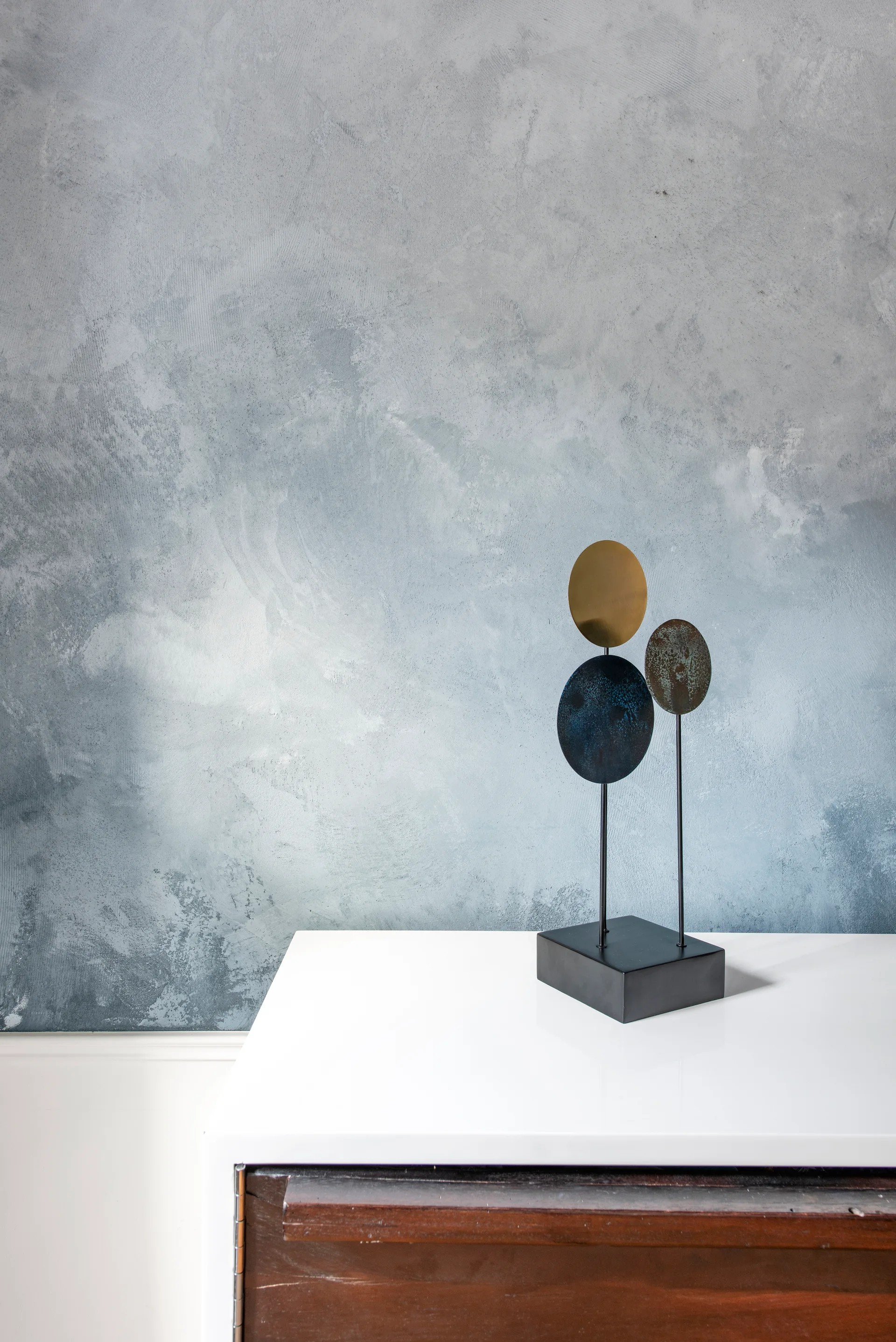 An abstract metal sculpture with circular discs rests on a glossy white console table against a textured blue-grey wall.