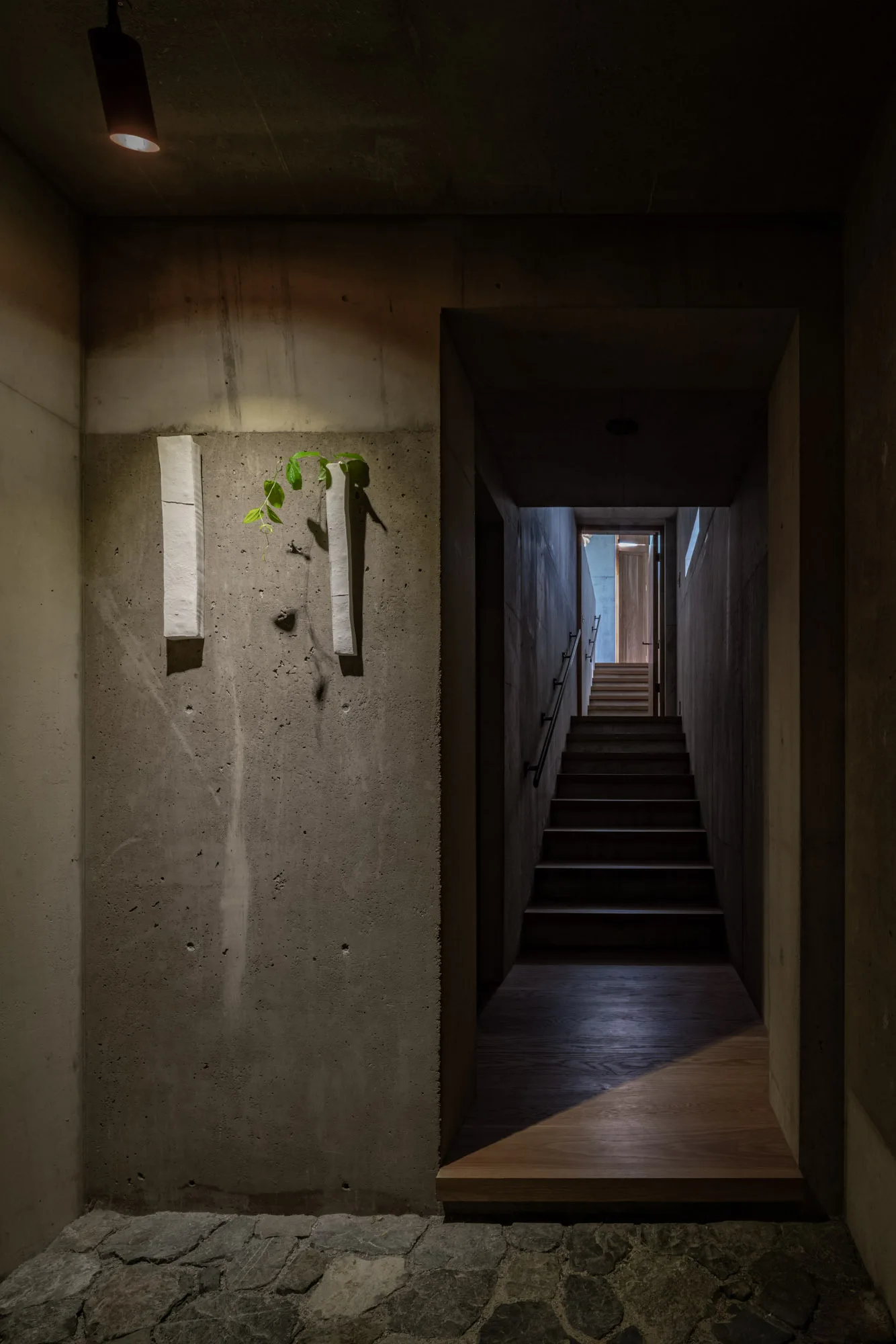 A dimly lit entryway featuring raw concrete walls, an irregular stone floor, two vertical white wall vases, and a dark wooden staircase.