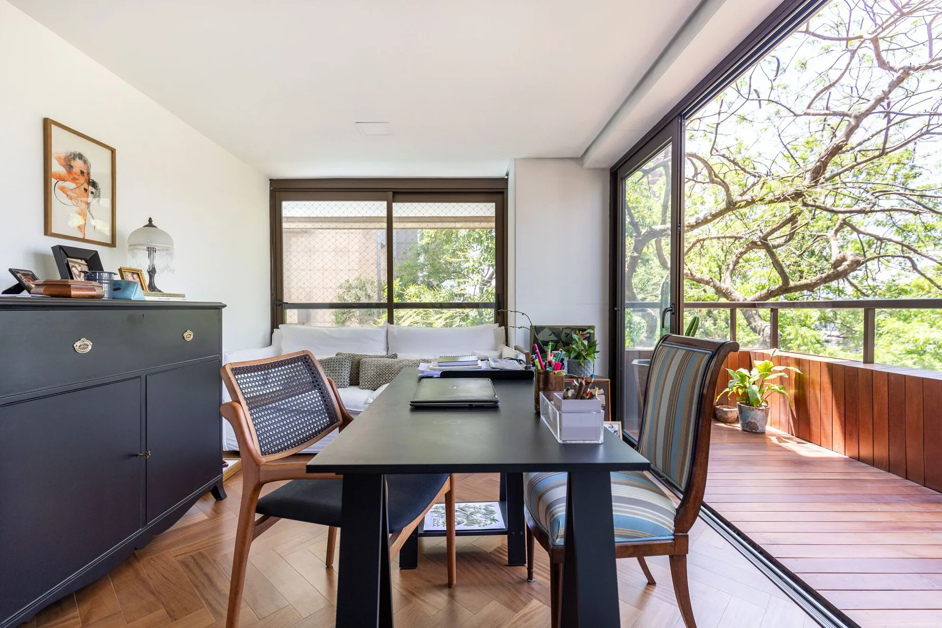 A bright home office featuring a black desk, mixed-style wood chairs, herringbone floors, and a sliding glass door opening to a wooden balcony.