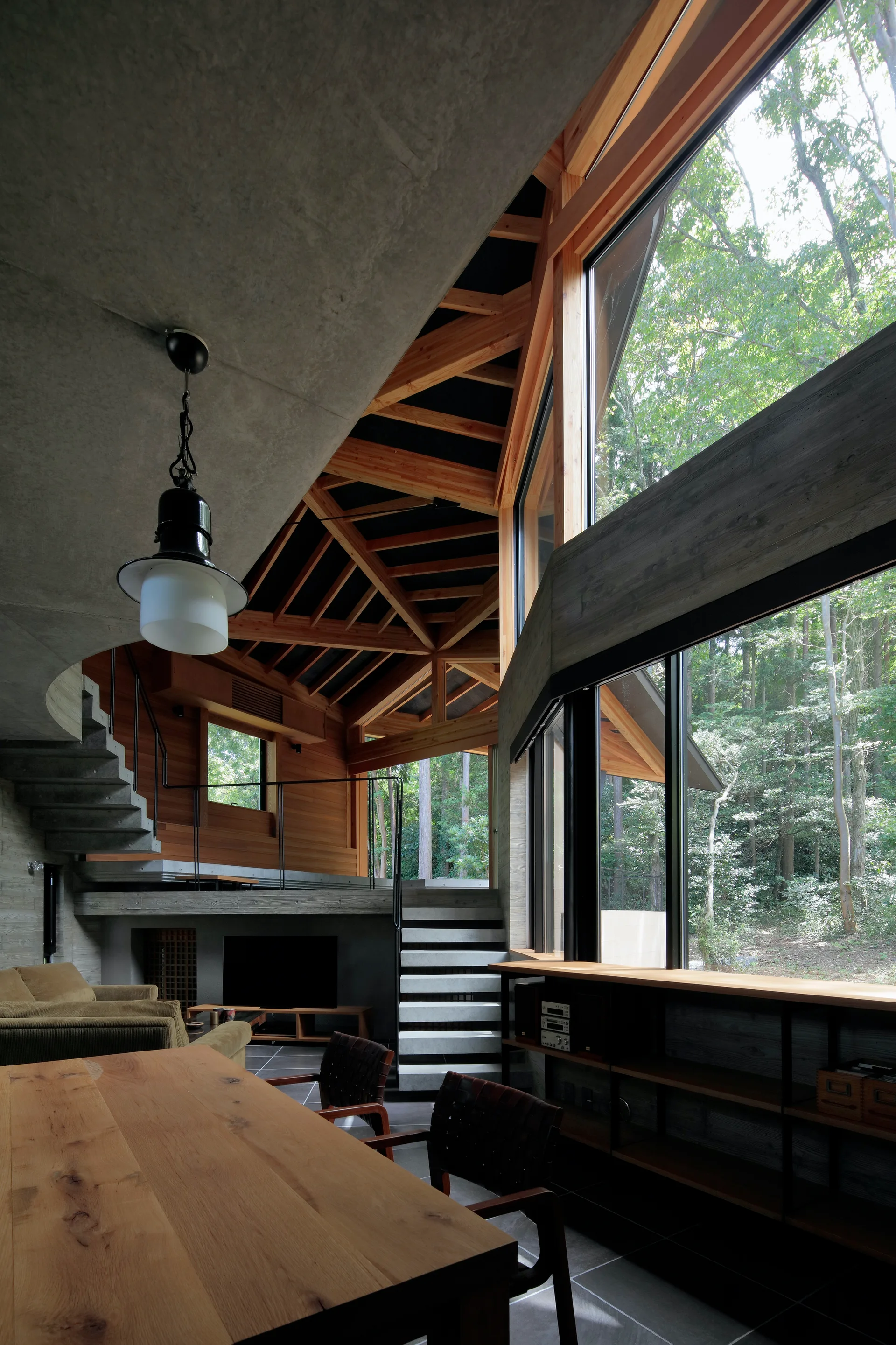 A multi-level modern interior featuring raw concrete walls, a geometric wooden rafter ceiling, and large windows looking out onto a dense forest.