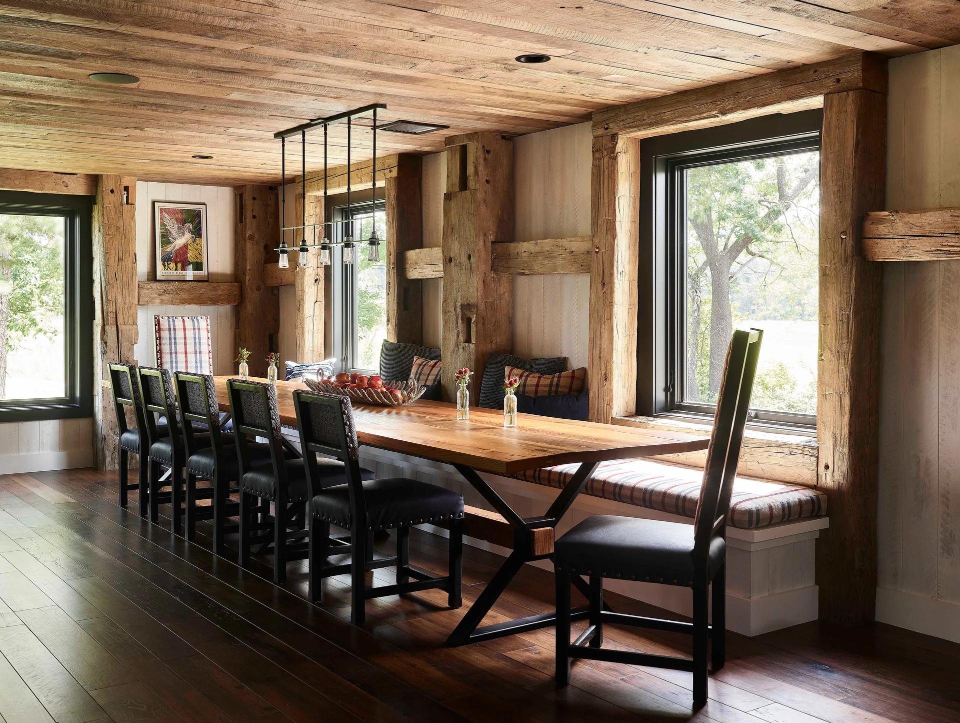 A rustic dining room features a long wooden table, dark upholstered chairs, and a built-in window bench beneath massive exposed timber framing.
