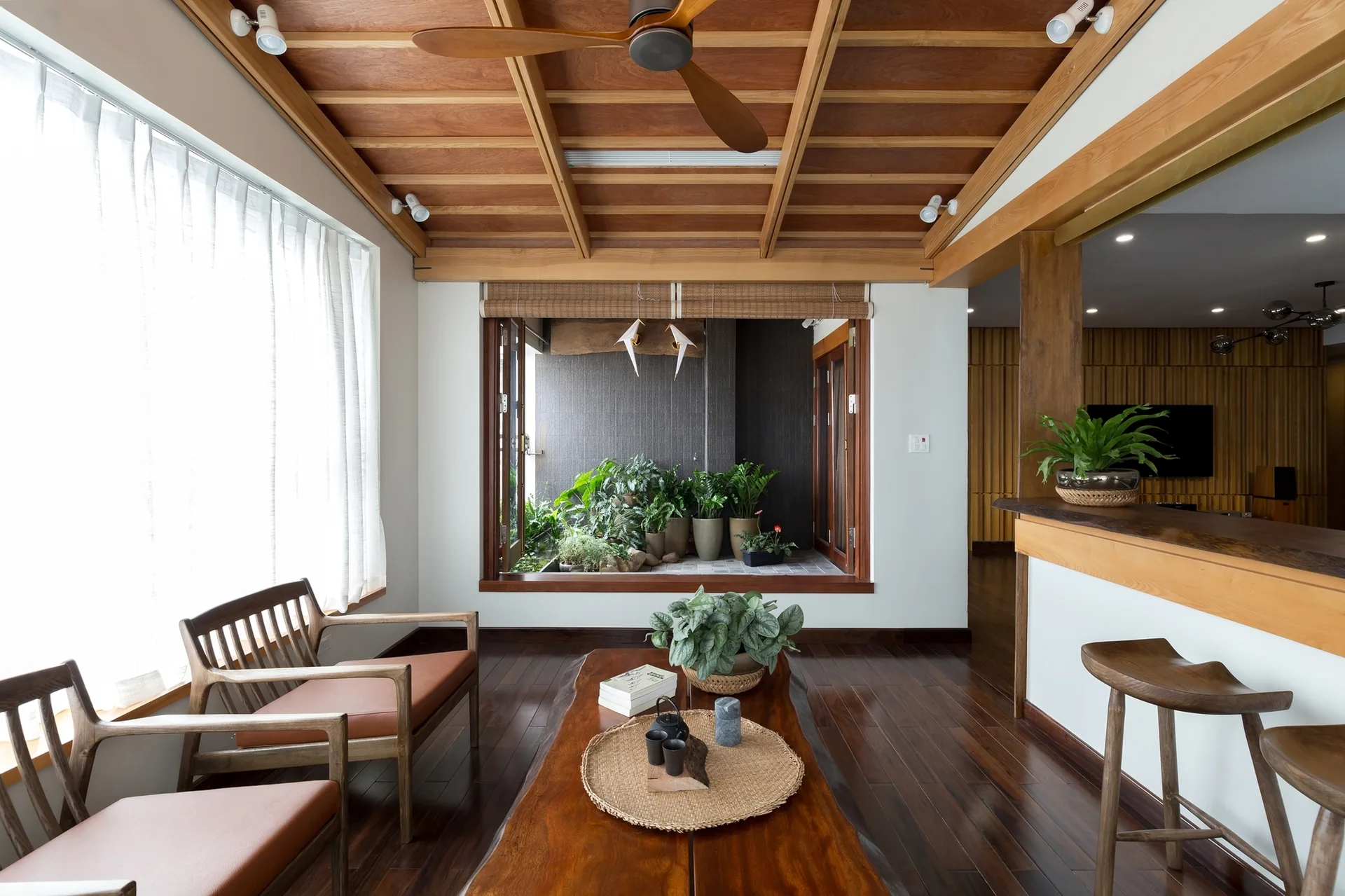 A modern living room features a warm wood ceiling with a fan, a live-edge coffee table, and a window viewing an indoor garden.