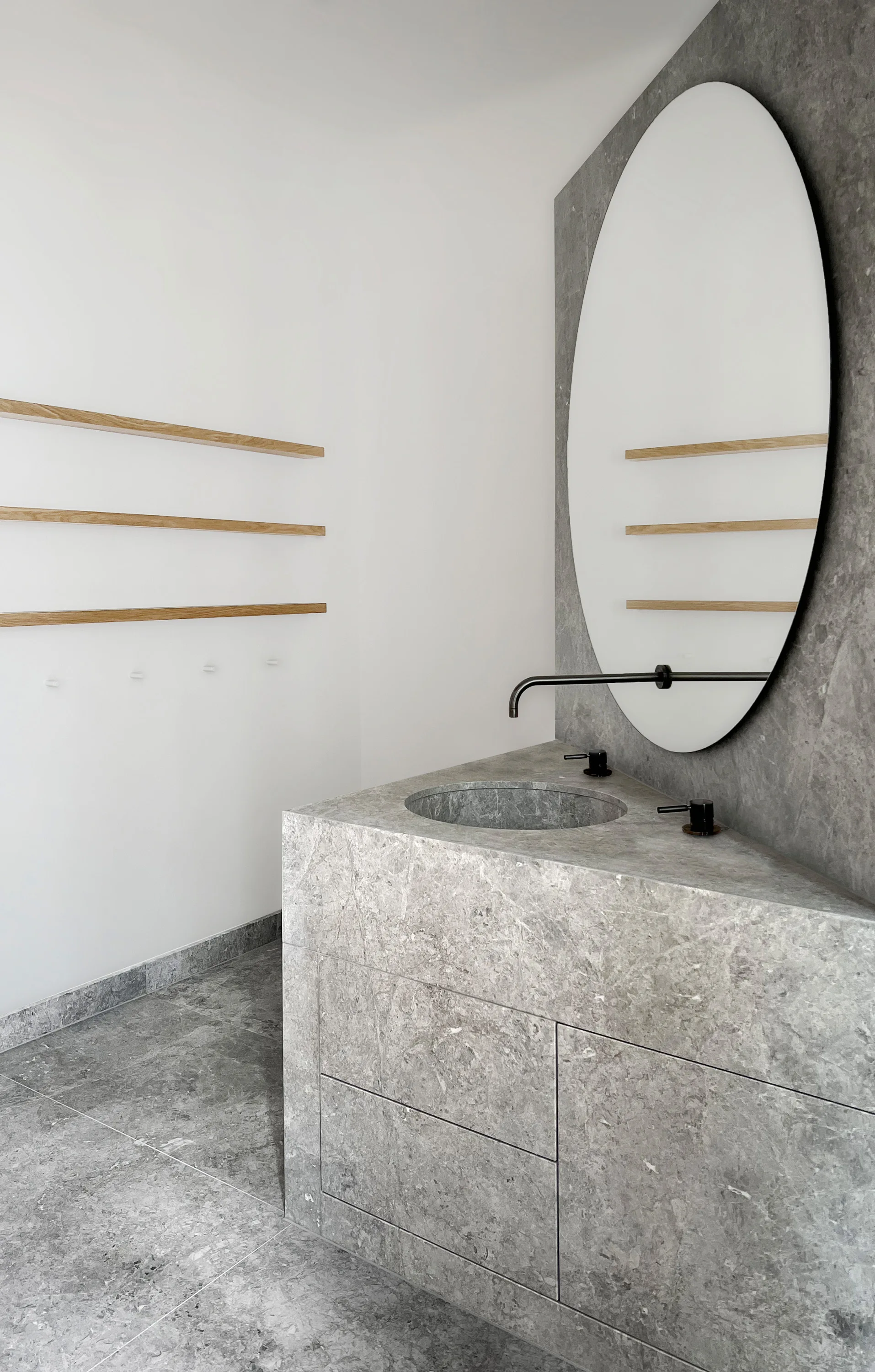 A minimalist bathroom featuring a grey stone vanity with an integrated sink, a large round mirror, and floating wood shelves on a white wall.