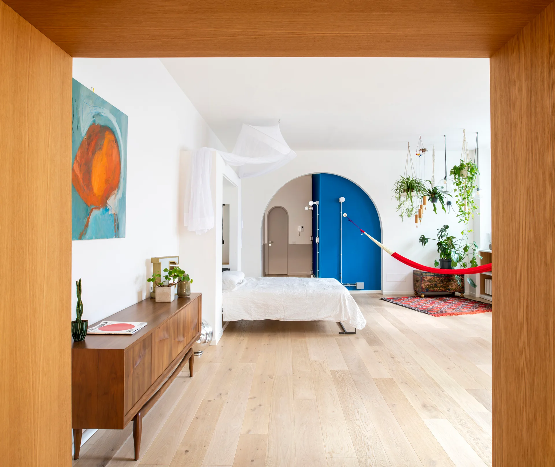 A view through a wooden door frame reveals a bright bedroom featuring a canopy bed, a blue painted archway, and a red hammock suspended over a patterned rug.