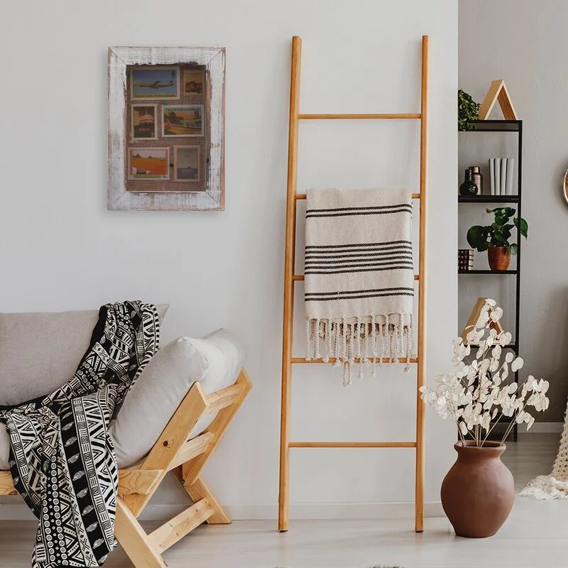 A minimalist living room featuring a wooden sofa with a patterned throw, a light wood blanket ladder, and a terracotta vase on the floor.