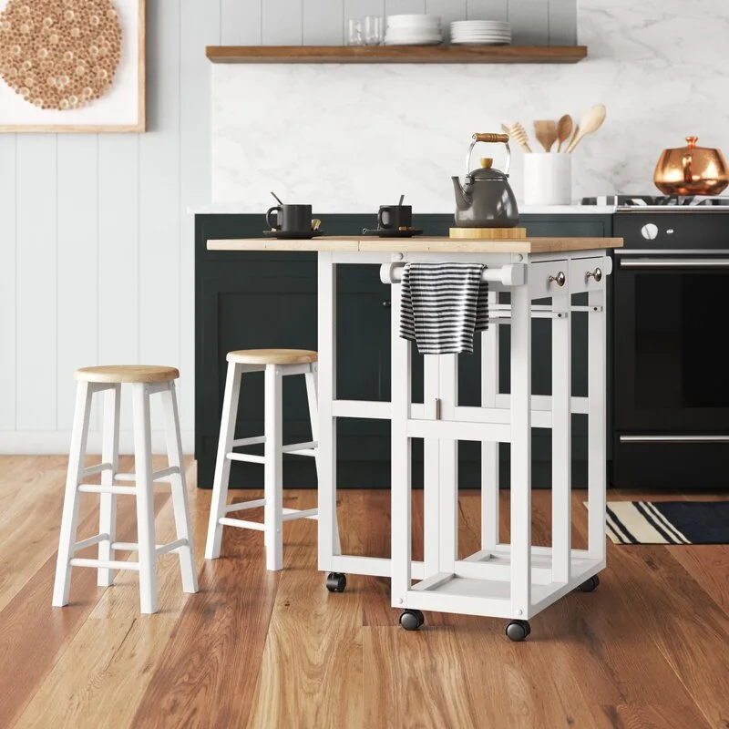 A white rolling kitchen island with a wood top and two matching bar stools stands in a kitchen with dark green cabinets and wood floors.