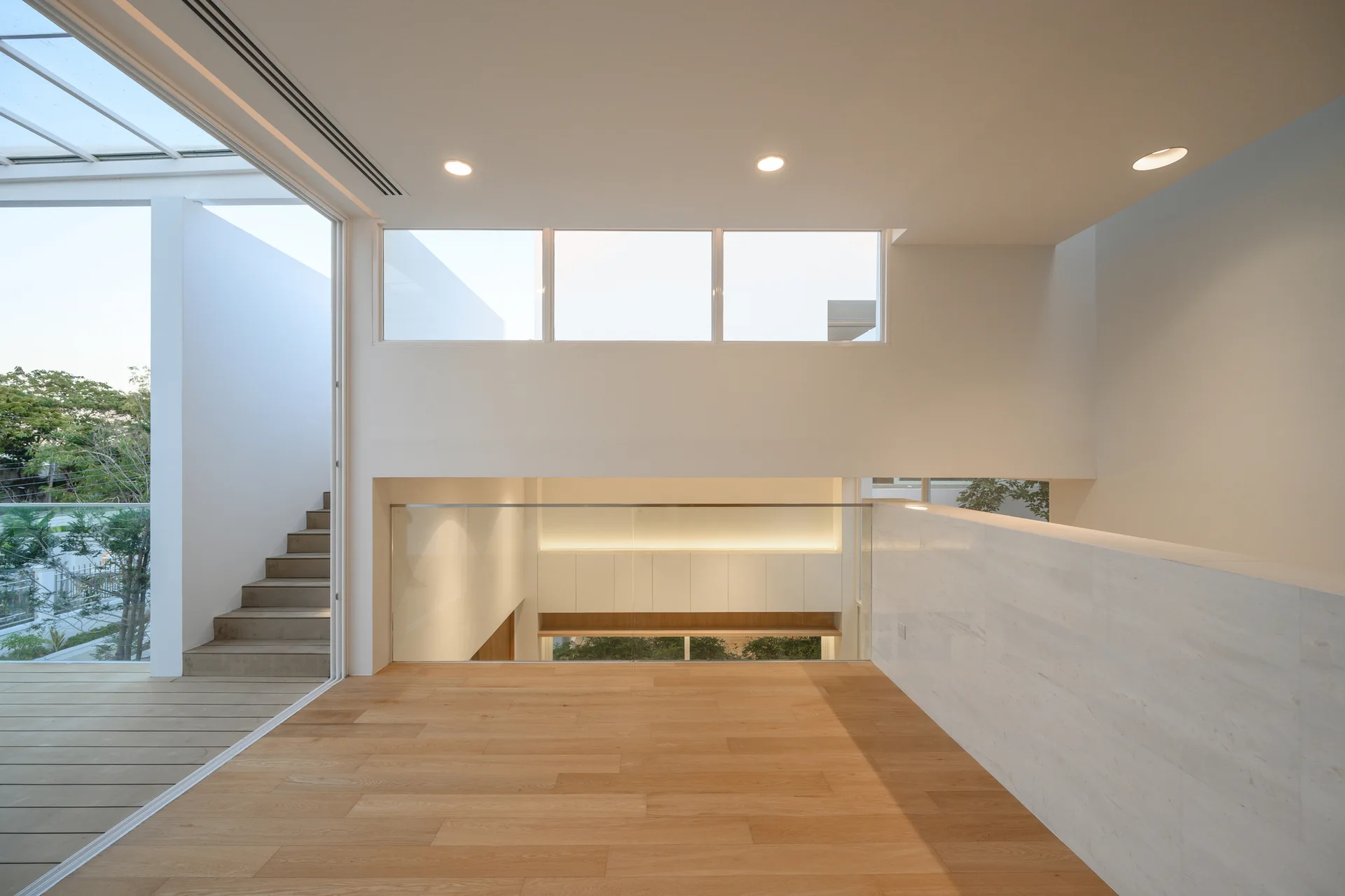 A modern minimalist landing features light wood floors, a transparent glass balustrade, and a view of lower-level cabinetry and high windows.