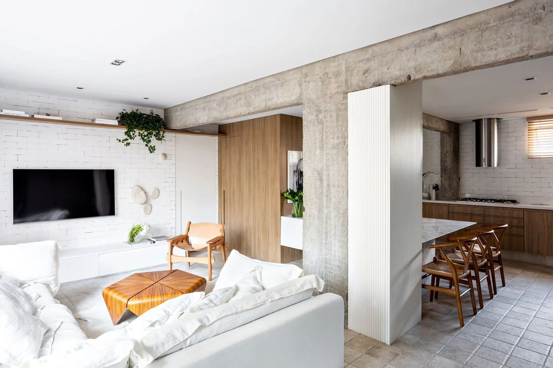 An open-concept living and dining space with an exposed concrete beam, white painted brick wall, white linen sofa, and wood dining chairs.
