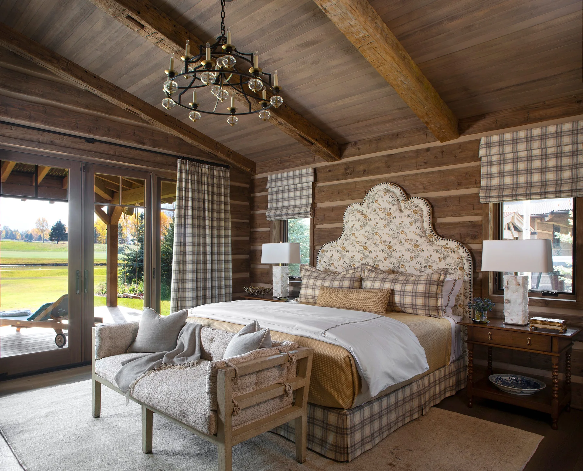 A rustic bedroom with exposed log walls, a wooden beamed ceiling, a floral headboard, plaid bedding, and an upholstered bench.