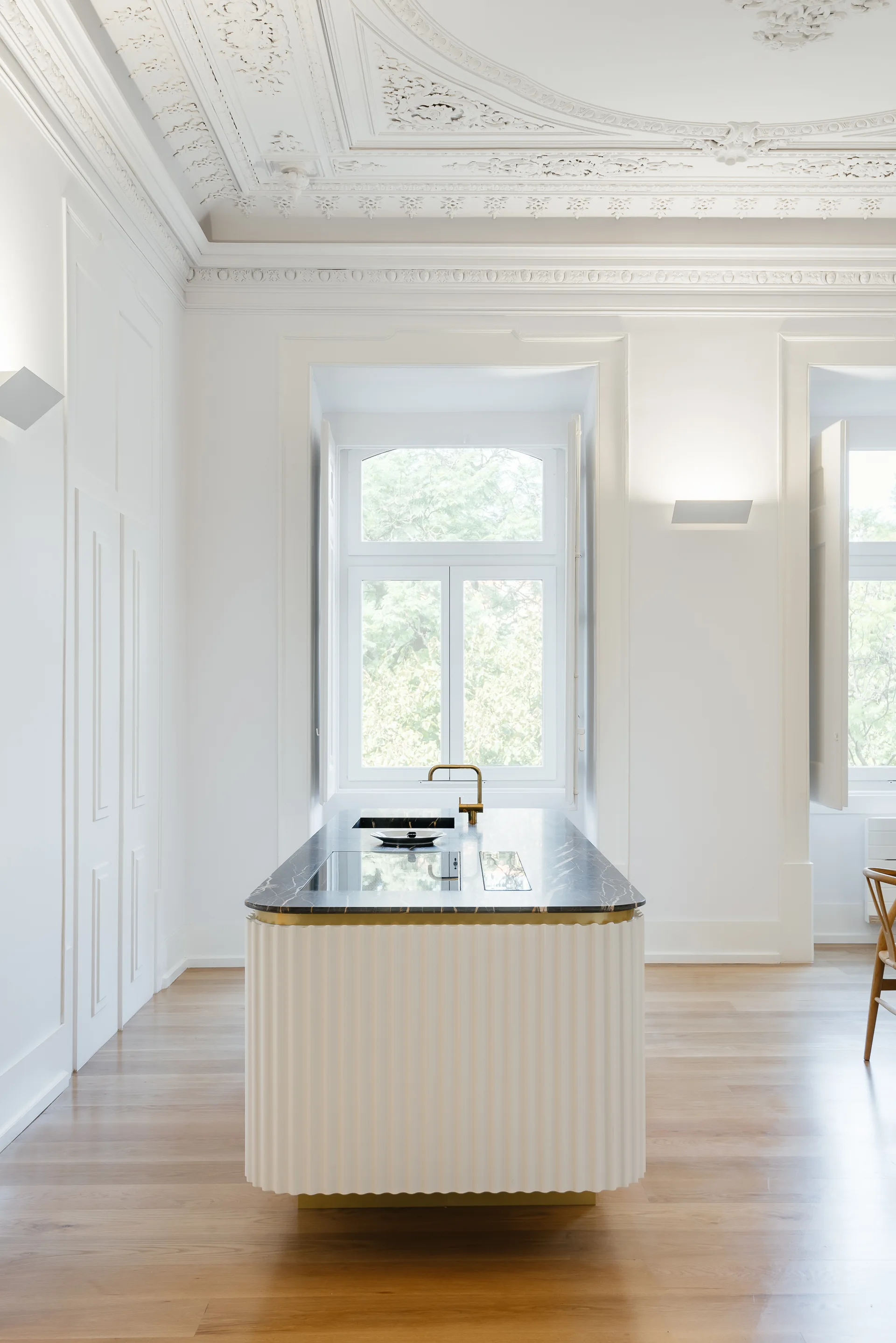A modern kitchen island with a fluted cream base and dark veined countertop stands on wood floors in a room with ornate white plaster ceiling moldings.