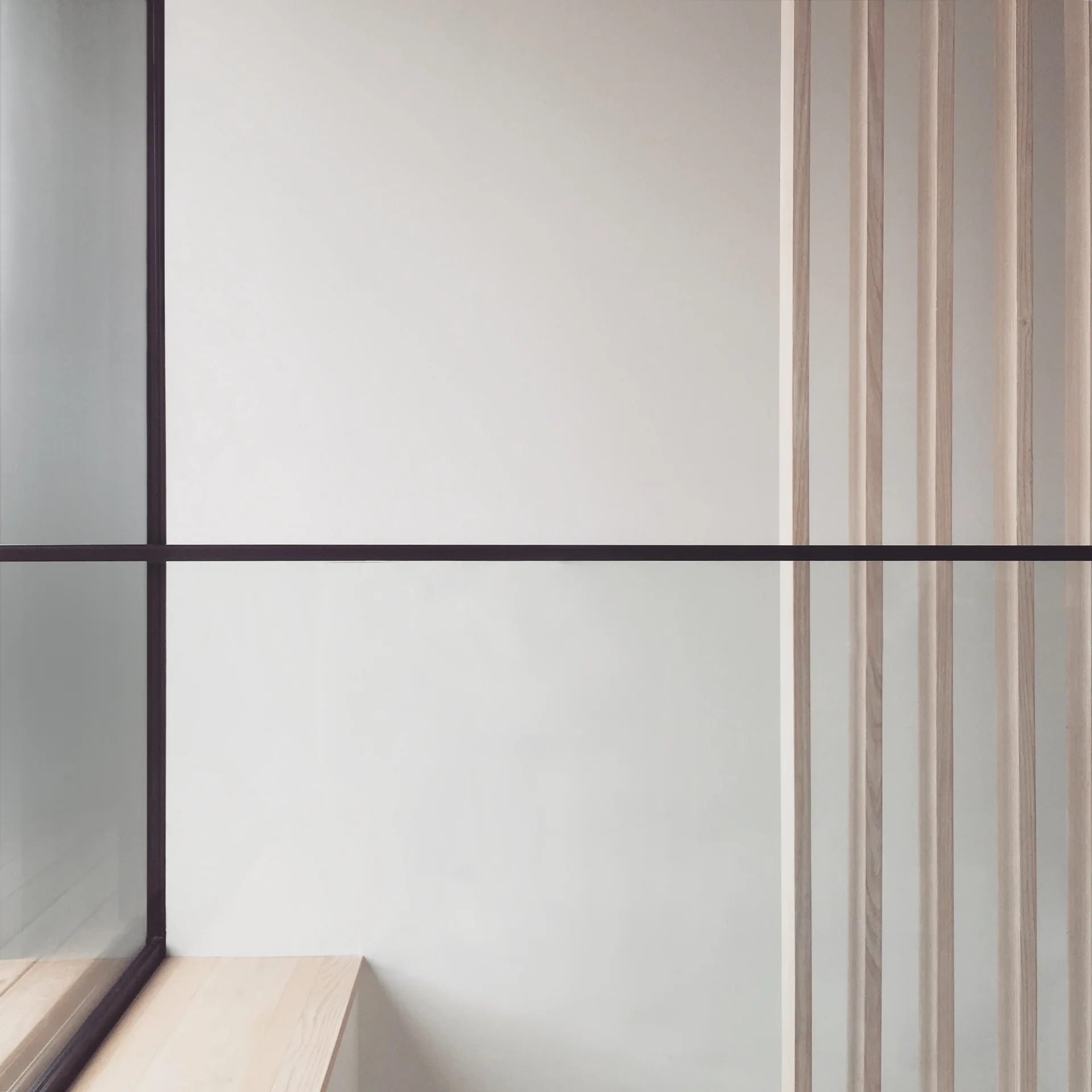 A minimalist interior wall with light wood vertical slats, a matching wood ledge, and a black metal-framed glass partition.