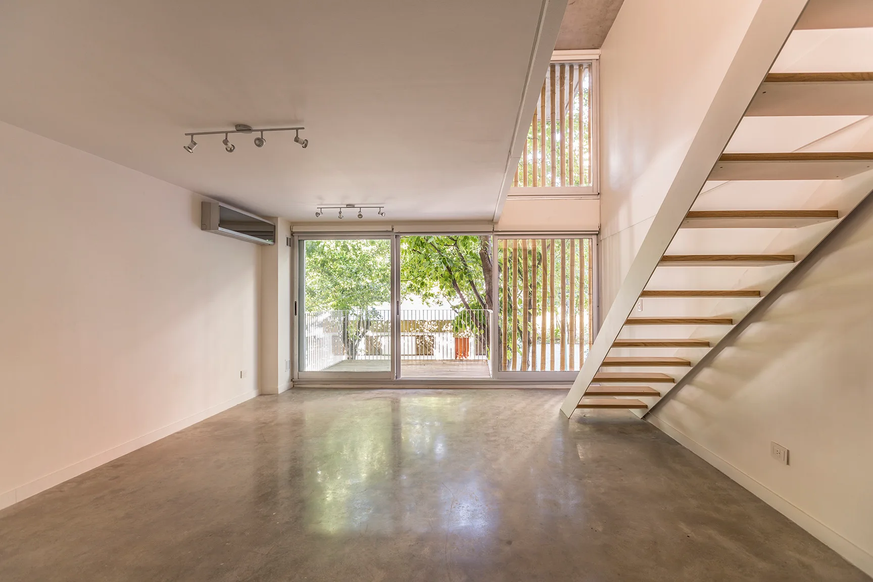An empty minimalist room features polished concrete floors, white walls, an open-riser staircase with wood treads, and large sliding glass doors.