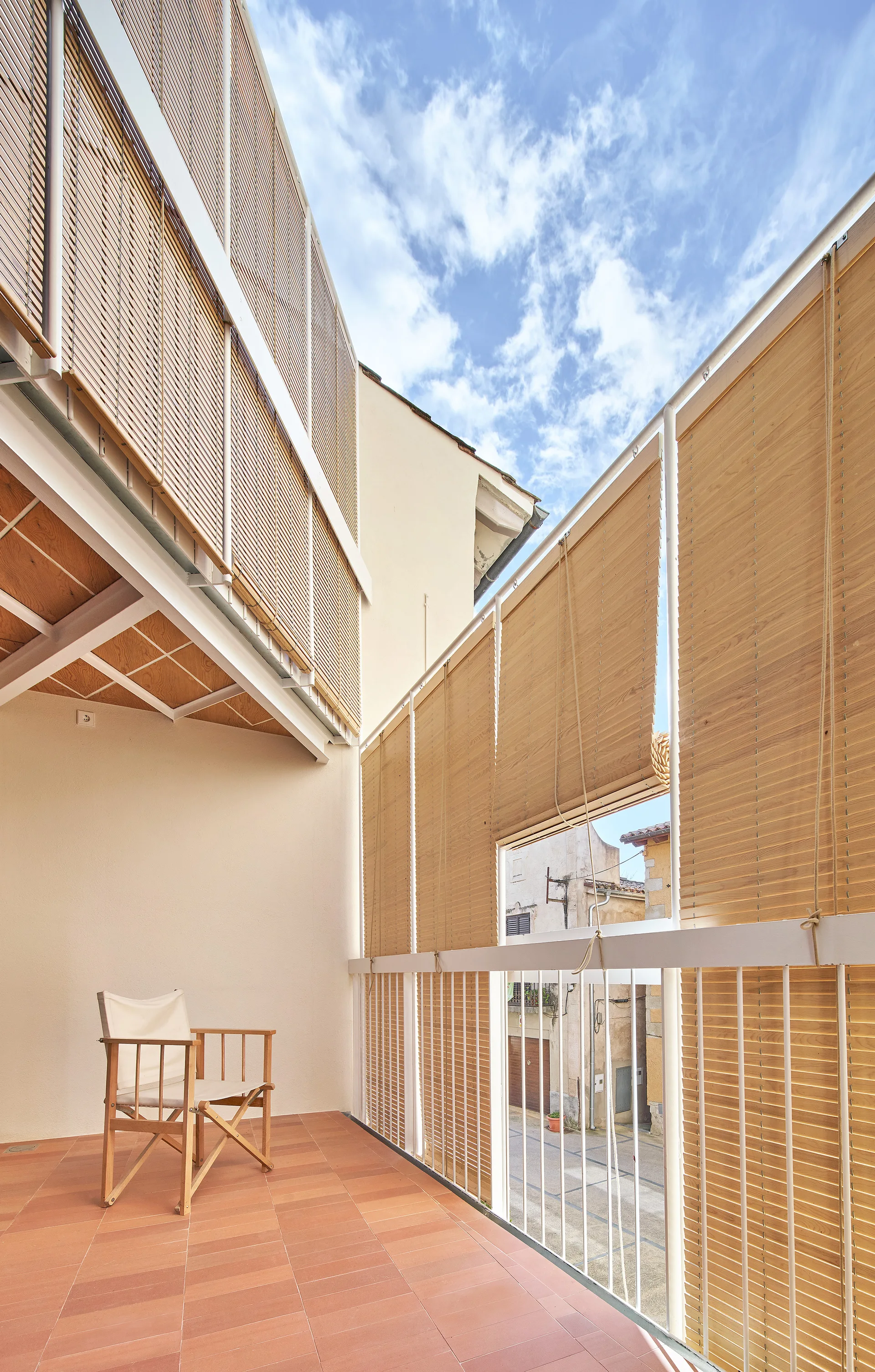 An outdoor balcony features terracotta tile flooring, a wooden folding chair, and roll-up bamboo privacy shades mounted on a white railing.