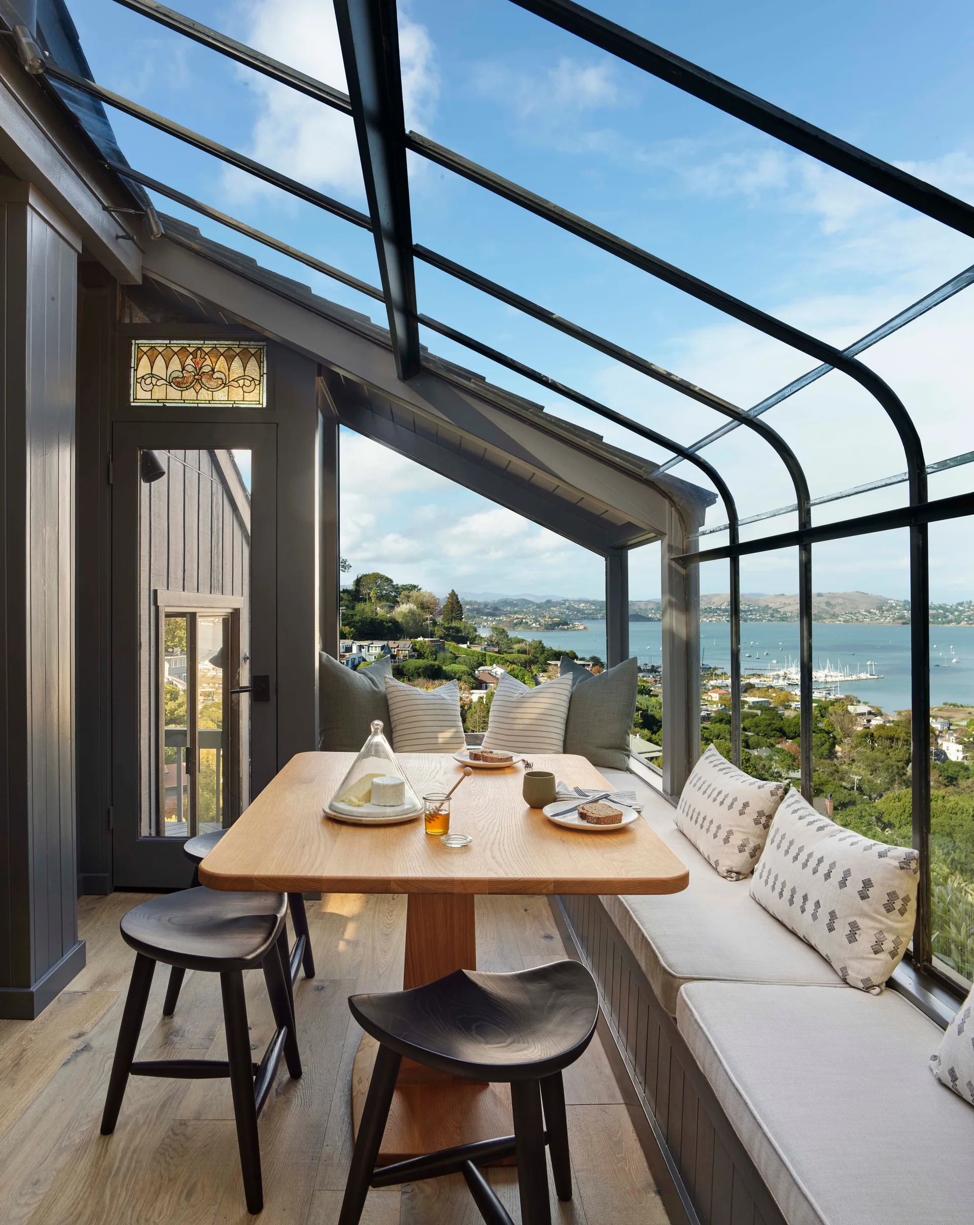 A sunroom dining nook with a wooden pedestal table, black stools, and a built-in window bench beneath a glass-panel roof overlooking a bay.