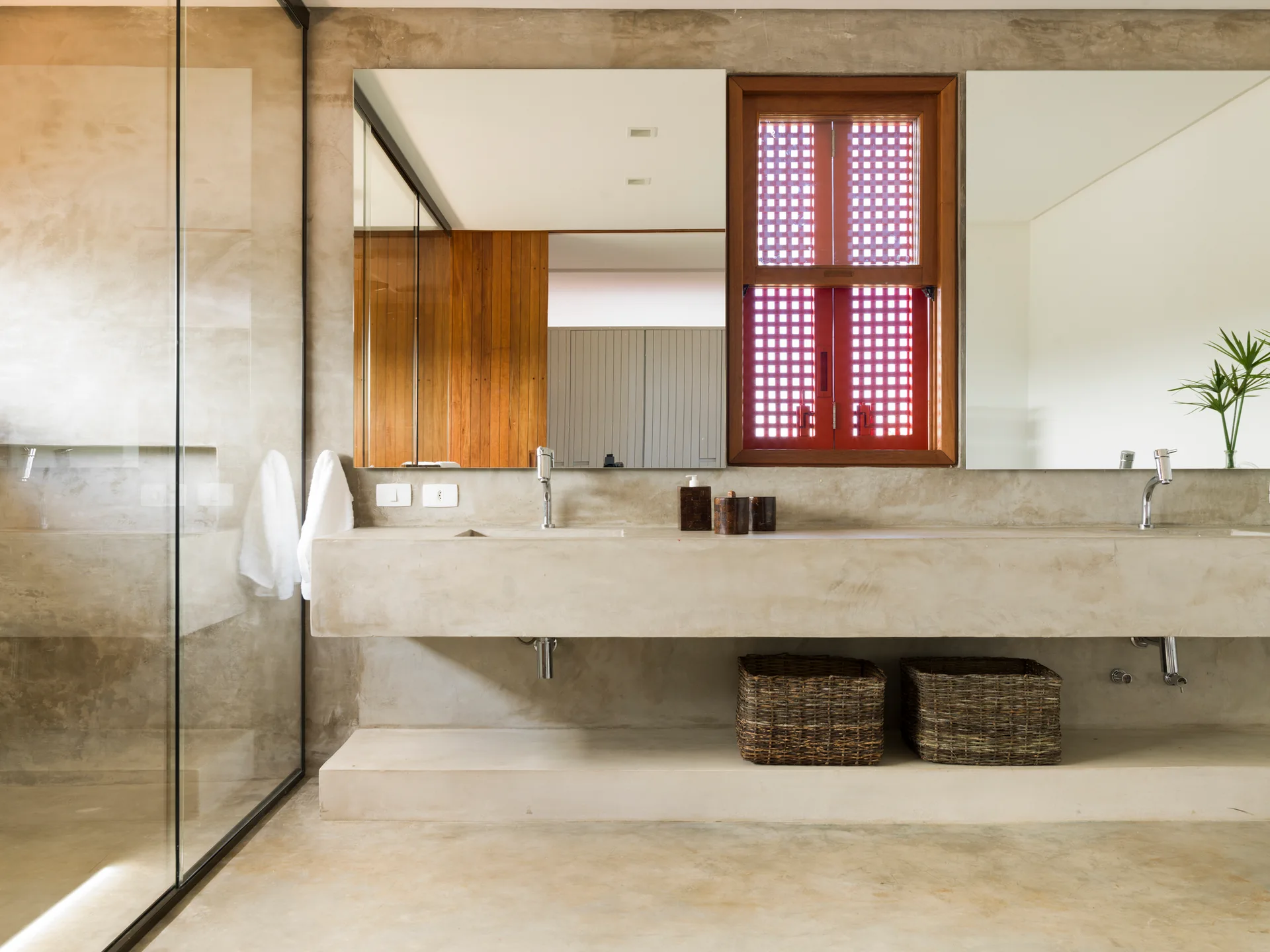 A minimalist bathroom featuring a floating concrete double vanity, two large wall mirrors, and a central red wooden window with a grid pattern.