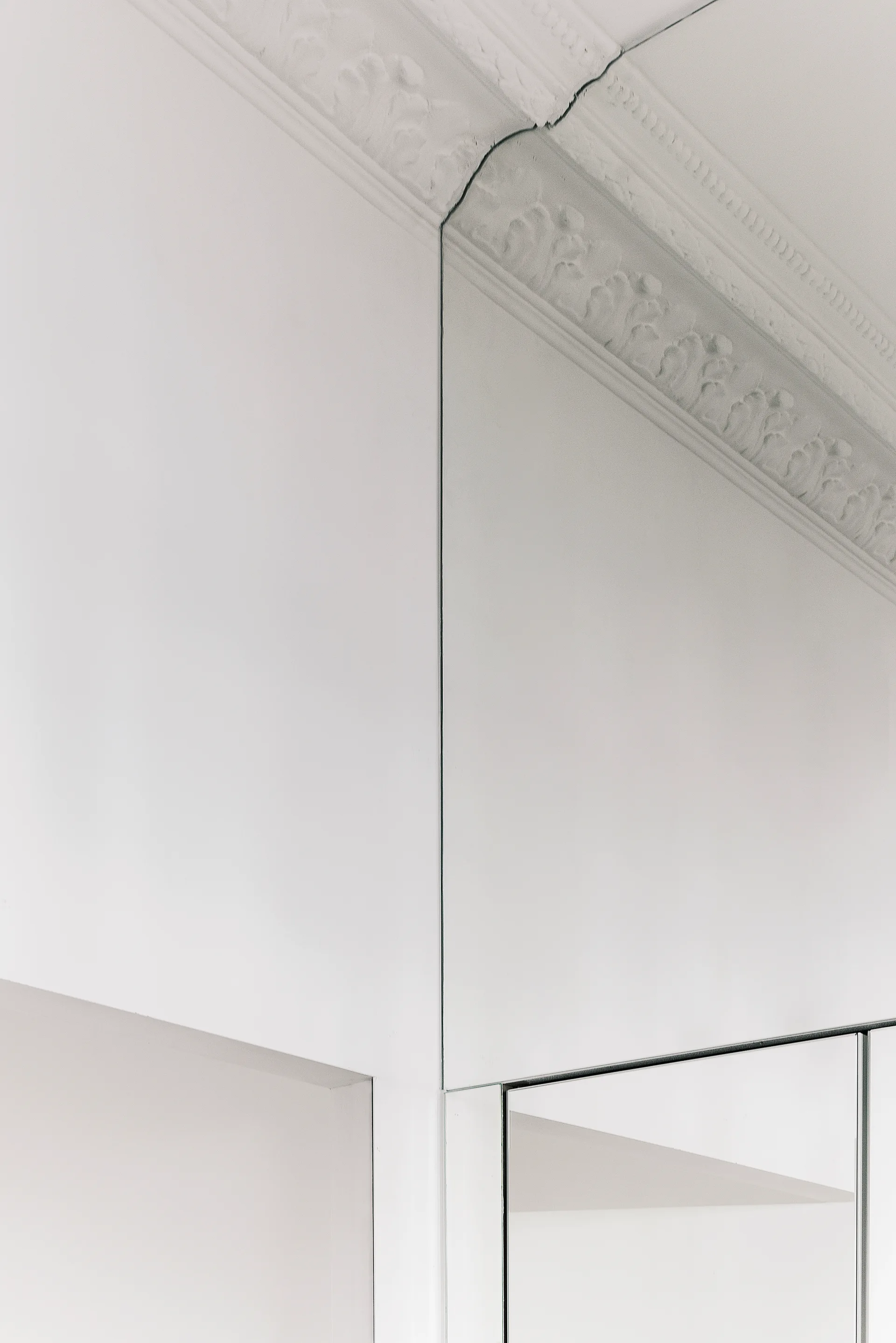 A close-up view of a room corner with ornate white ceiling molding meeting a flat white wall and a vertical mirrored wall panel.