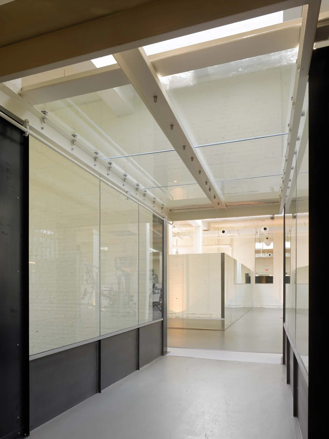 An industrial hallway featuring glass partition walls, a skylight ceiling with white beams, and painted white brick walls.