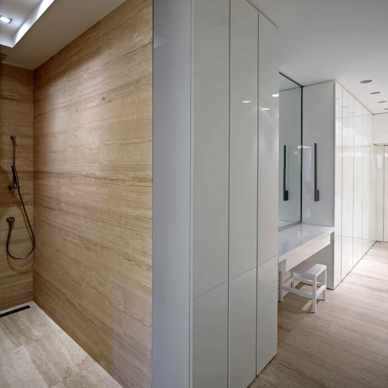 A modern bathroom featuring beige stone wall tiles in the shower area next to high-gloss white built-in storage cabinets and a sleek vanity desk.