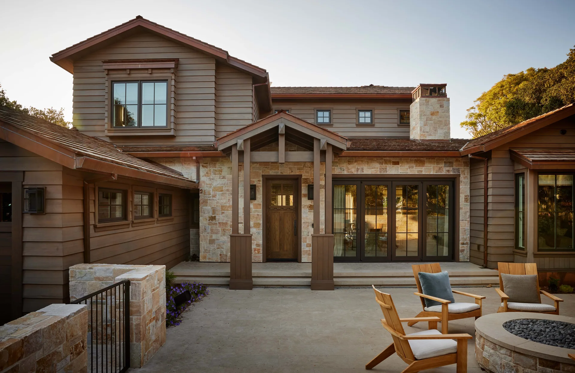 The exterior of a craftsman-style home featuring wood siding, a stone facade, and a stone patio with a round fire pit and wooden armchairs.