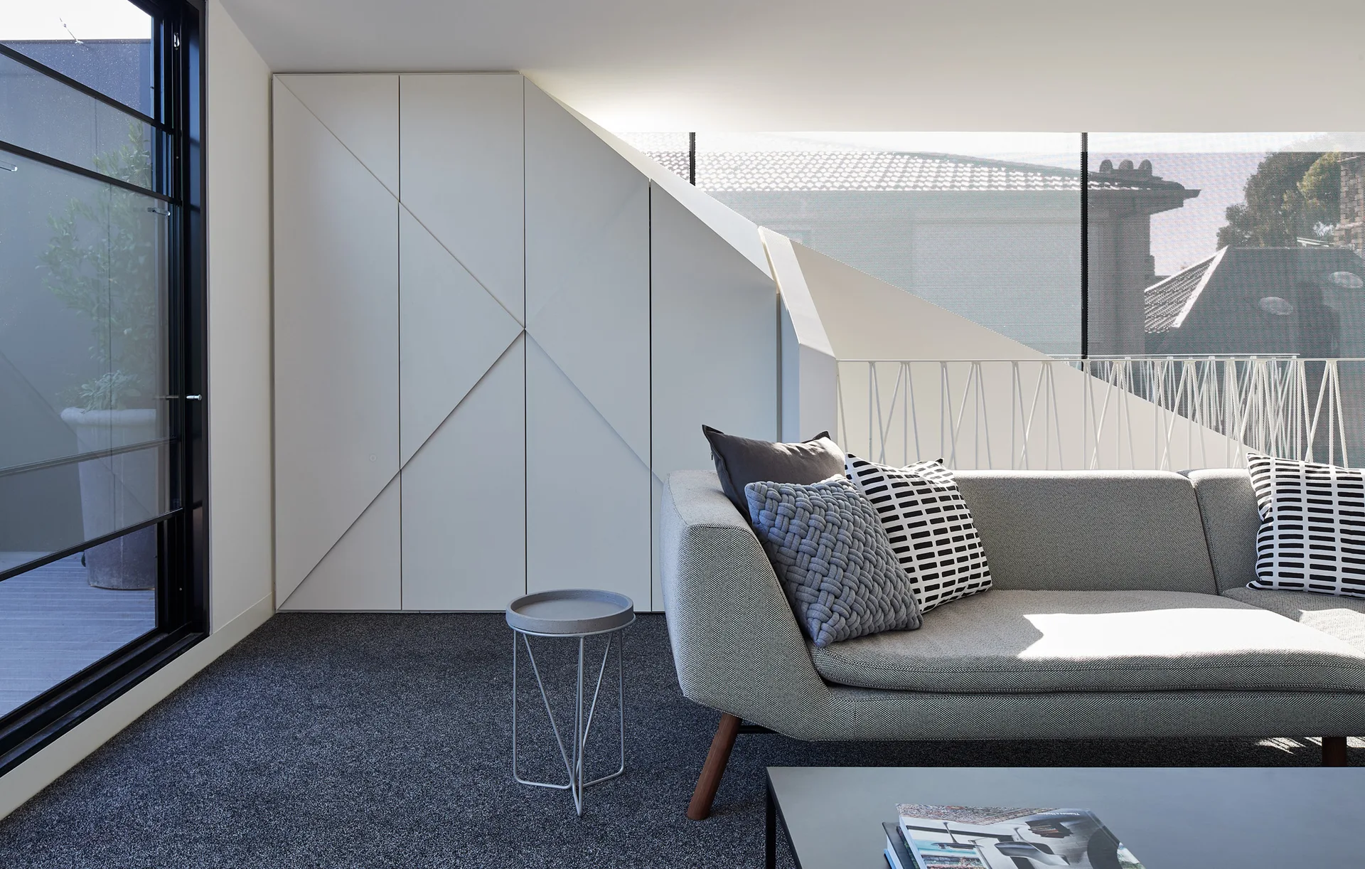 A modern living room features a gray fabric sofa with patterned pillows, a round metal side table, and a white built-in cabinet with geometric details.