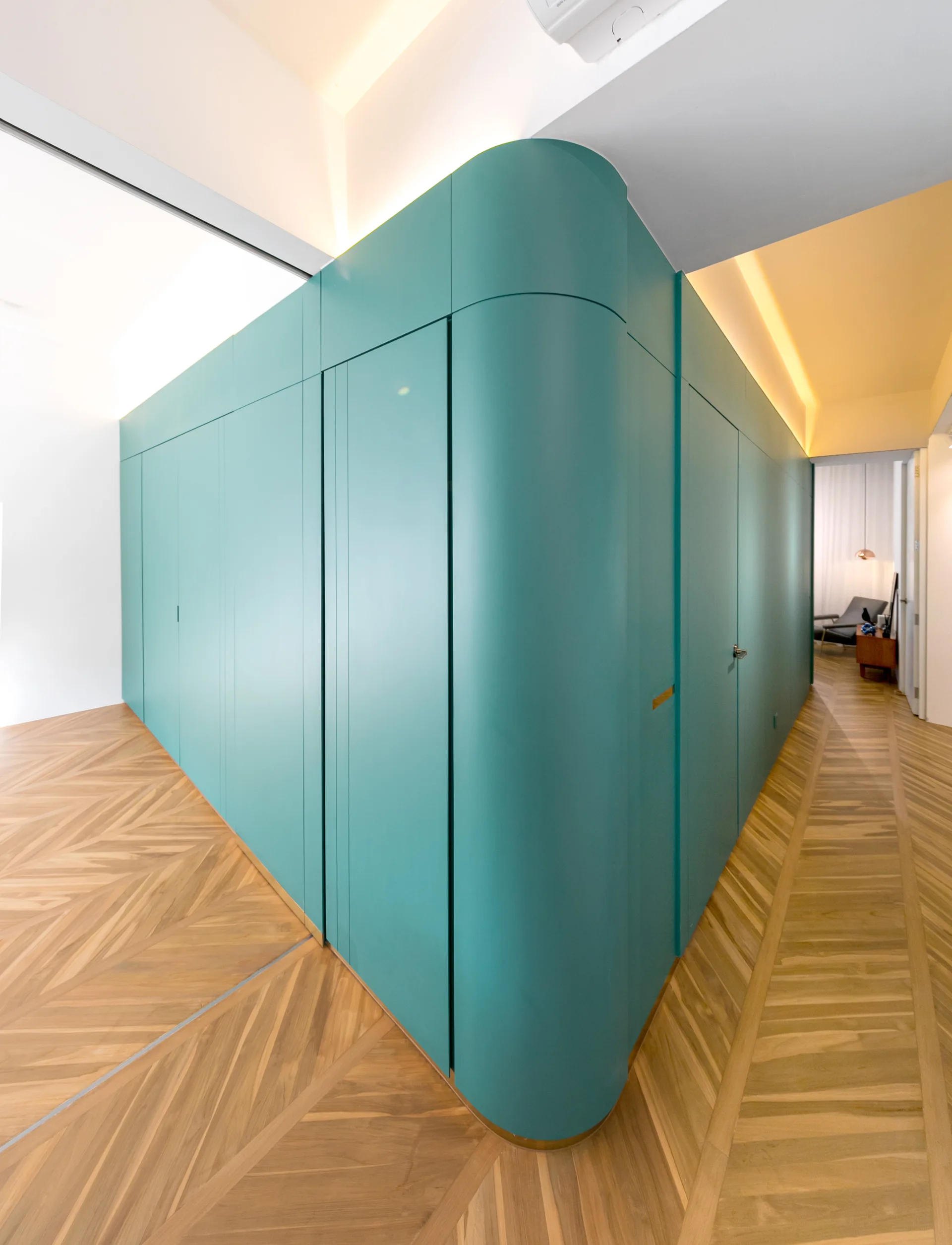A curved teal built-in cabinetry unit stands in a hallway on light brown herringbone wood flooring beneath warm cove lighting.