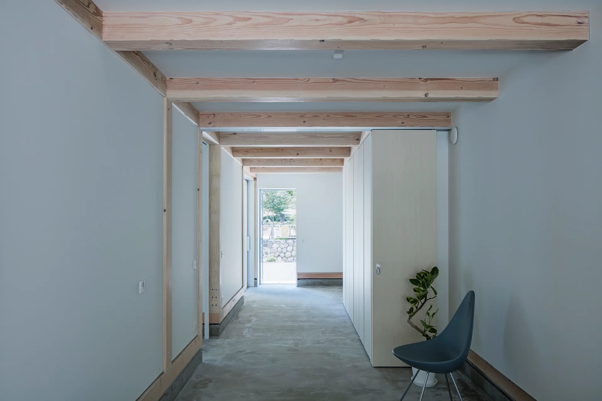 A minimalist hallway features exposed light wood ceiling beams, a smooth concrete floor, and built-in cabinets leading to an outdoor doorway.