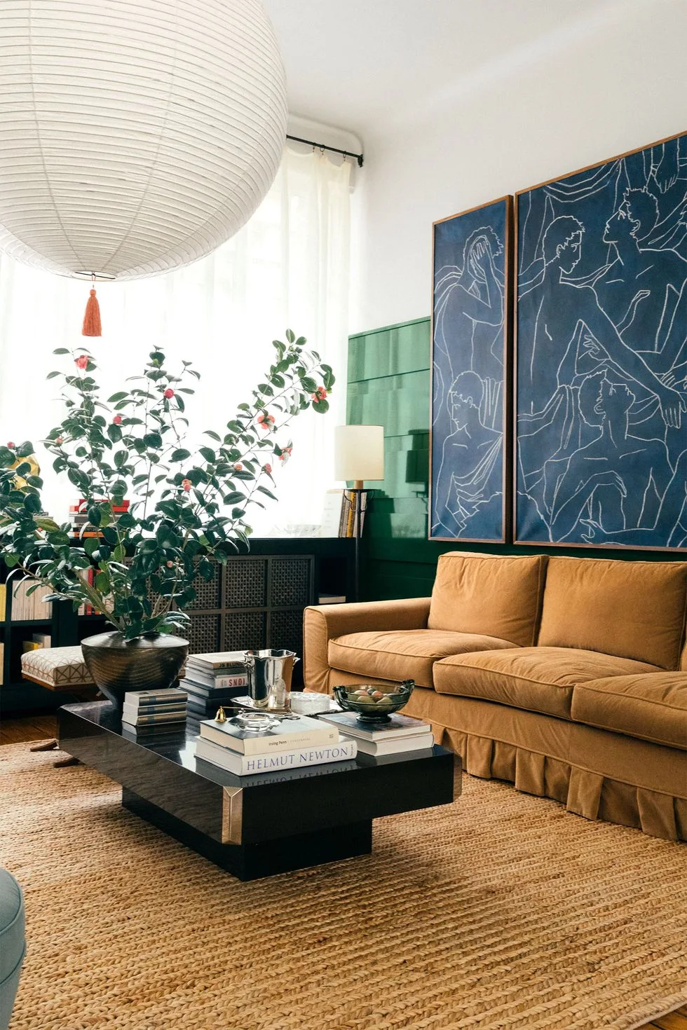 An eclectic living room features an ochre velvet skirted sofa, a black lacquer coffee table, a jute rug, and glossy green wall panels with blue art.