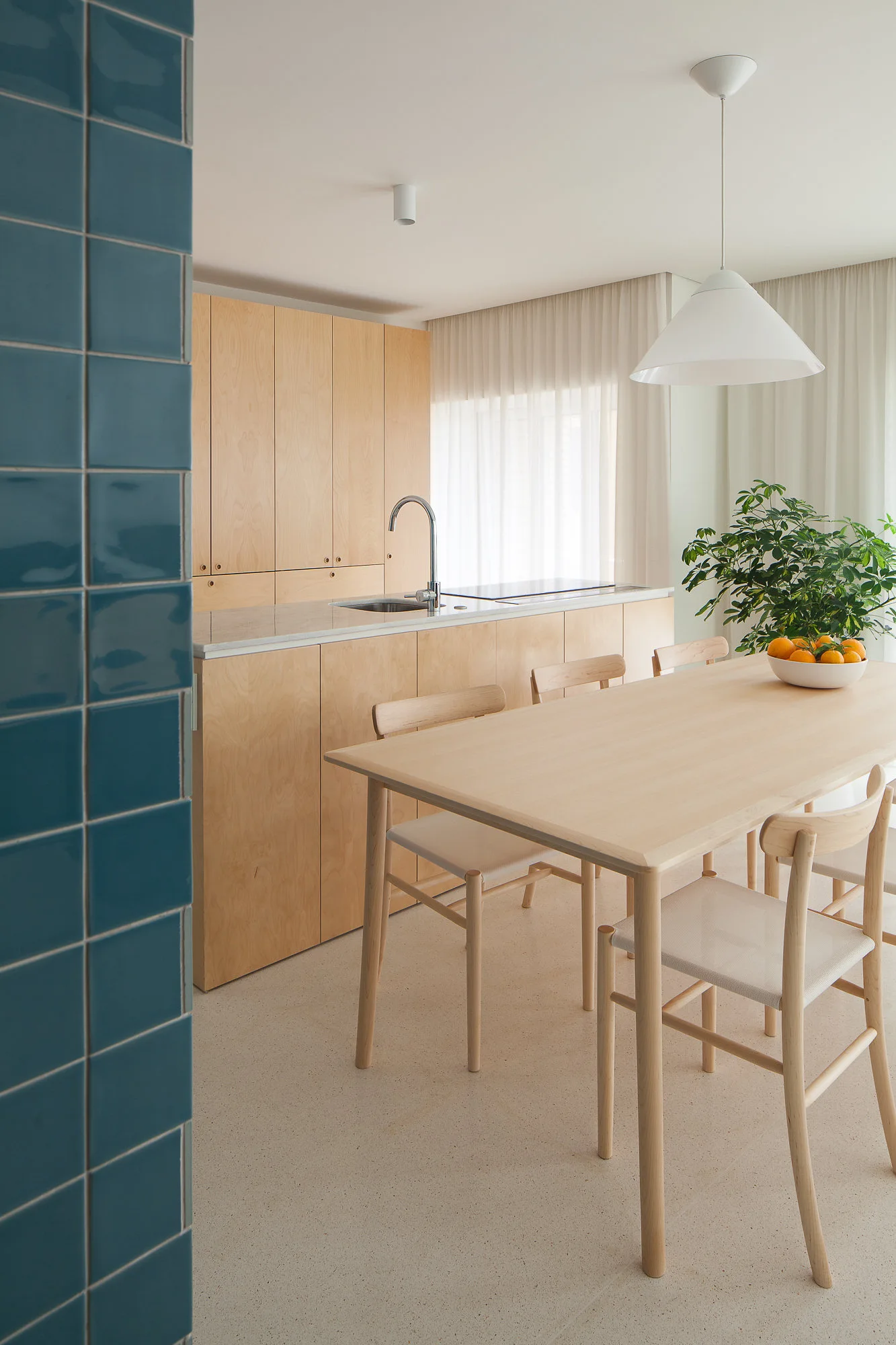 A minimalist kitchen features light wood cabinetry, a matching dining table, a white pendant lamp, and a glossy teal tiled pillar.