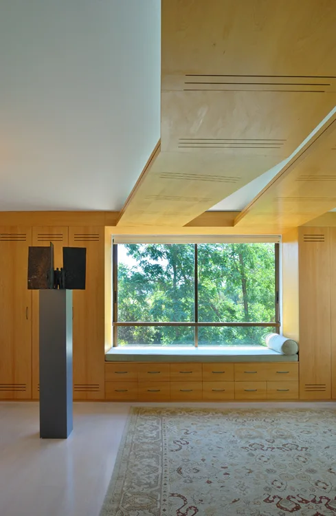 A light wood built-in window seat is flanked by matching cabinets under a wood ceiling panel, with a patterned rug and dark sculpture in the room.