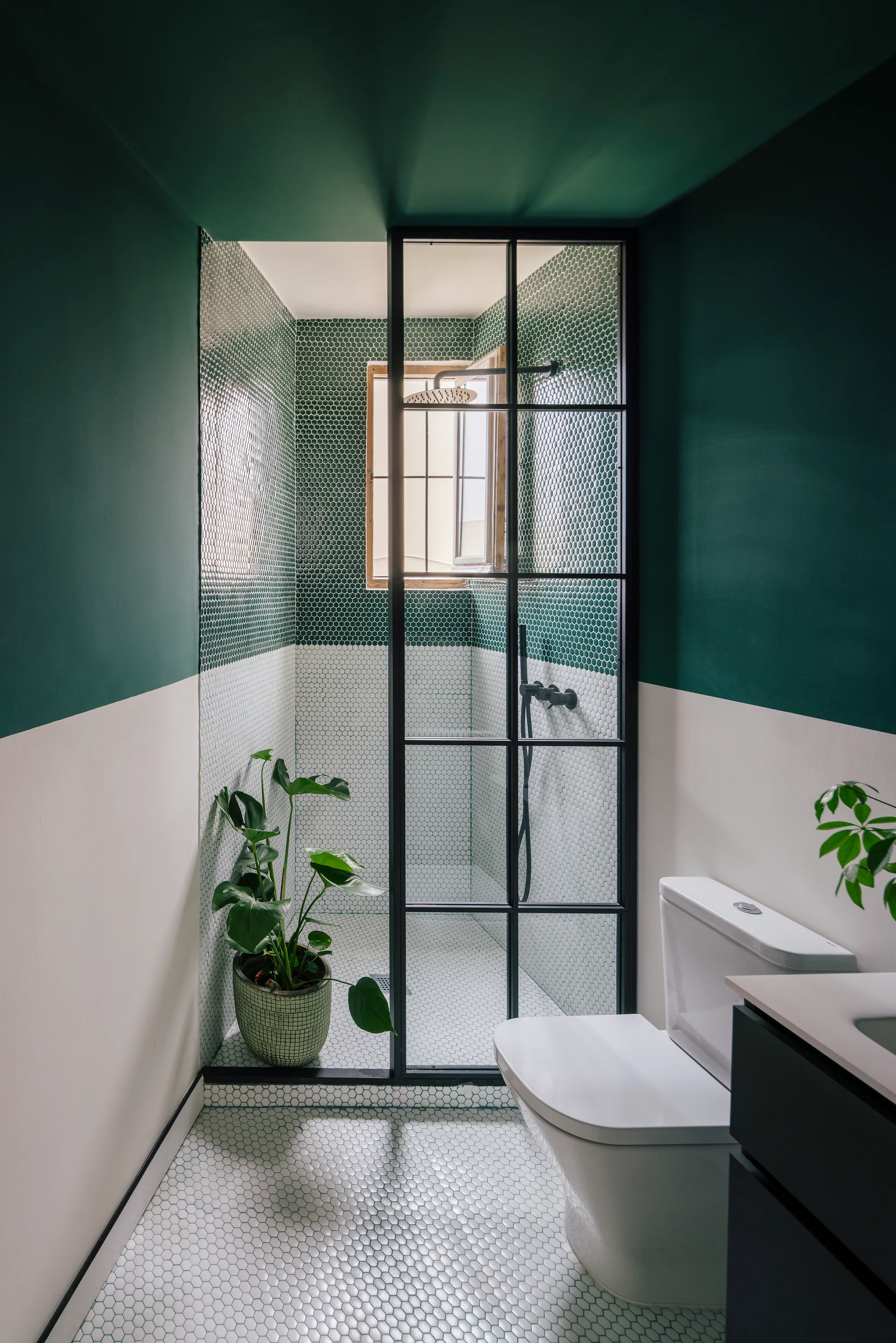 A modern bathroom features a split green and white wall, a walk-in shower with a black grid glass door, and white penny tile floors.