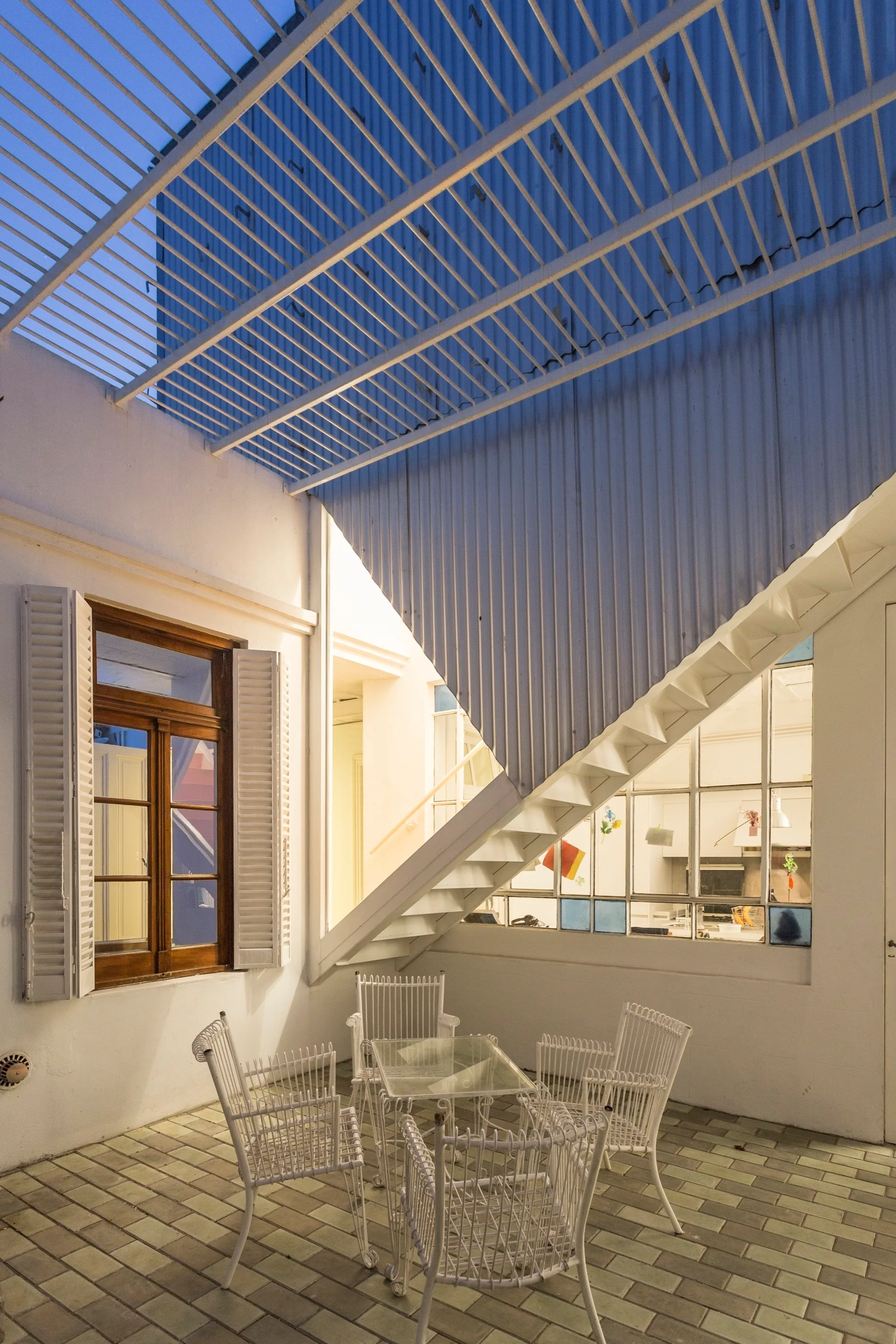 A white-walled courtyard patio features white metal wire chairs around a glass-top table beneath a diagonal staircase and slatted metal canopy.