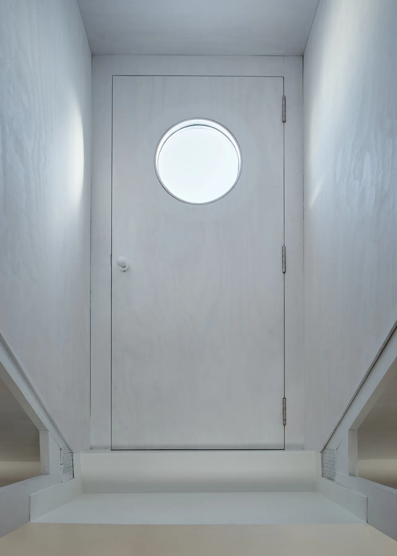 A minimalist white wooden door with a circular porthole window sits at the top of a simple white staircase under diffused lighting.