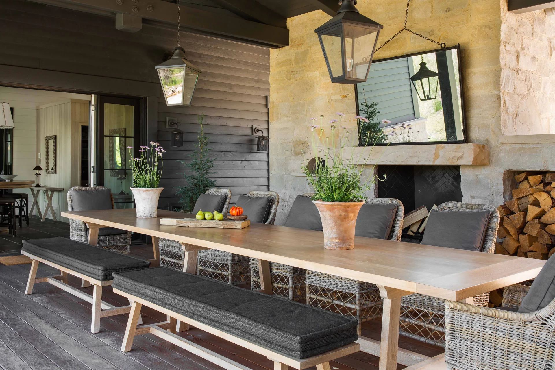 A covered outdoor patio features a long wooden dining table with grey-cushioned benches and wicker chairs in front of a stone fireplace.