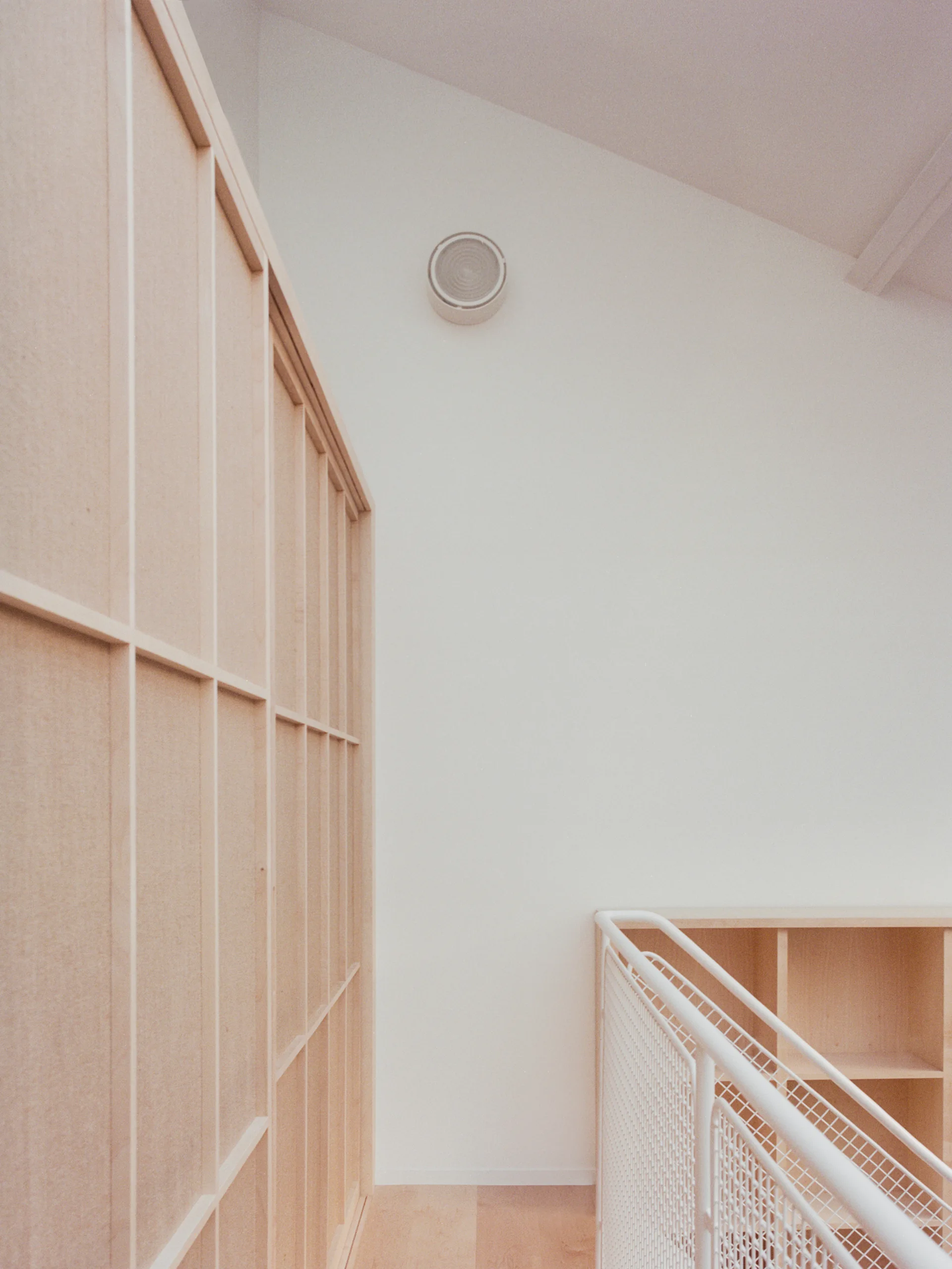 A light wood grid-paneled wall stands next to a white metal mesh railing on a bright landing under a sloped ceiling.