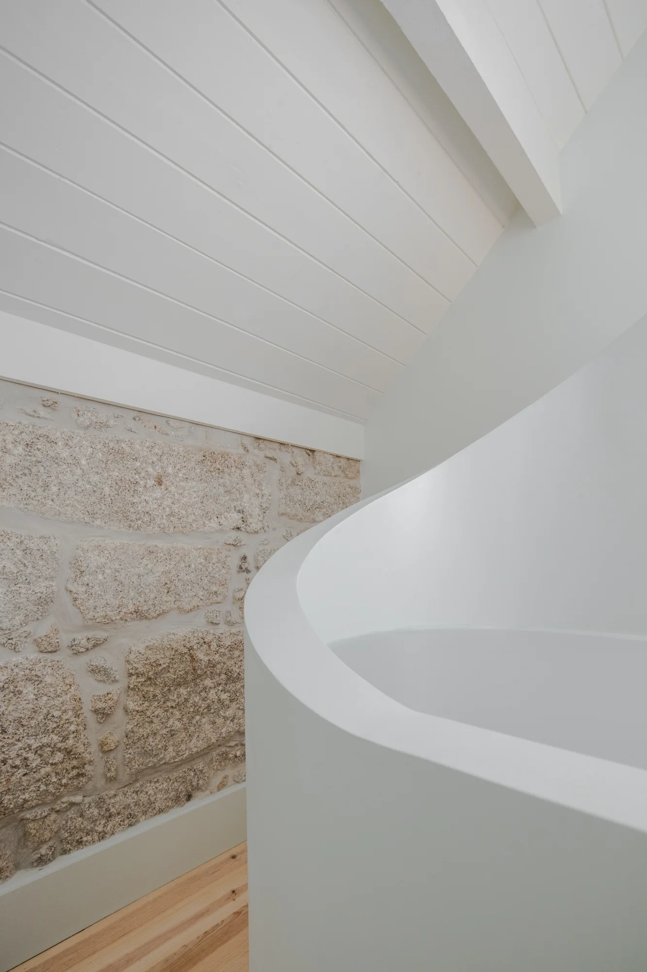 A close-up of a white curved bathtub set against a textured light grey stone wall and under a white sloped plank ceiling.