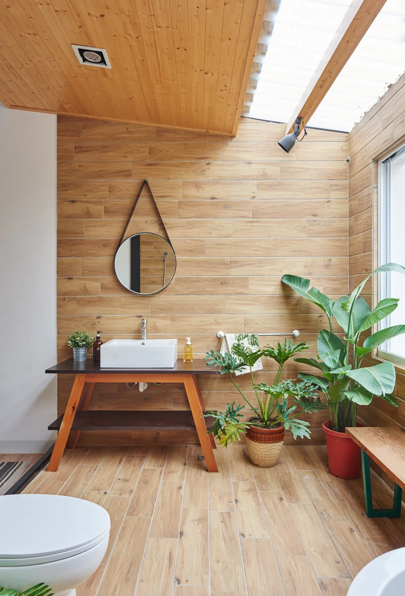 A bathroom features wood-plank walls and flooring, a wooden vanity with a vessel sink, a hanging round mirror, and potted plants under a skylight.