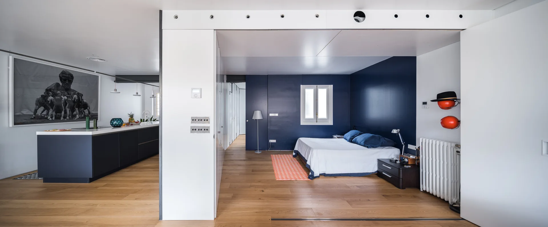 A modern open-concept apartment interior featuring a bedroom with a navy blue accent wall next to a kitchen with a dark blue island.