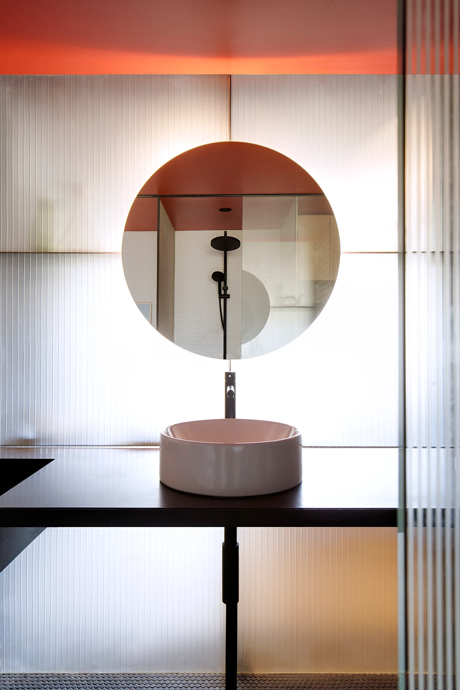 A minimalist bathroom vanity with a round white vessel sink and circular mirror in front of a translucent fluted glass partition.